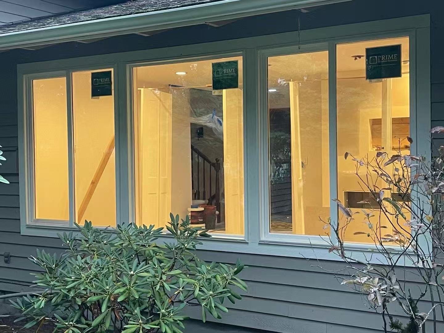 Exterior of a house with large front windows showing the illuminated interior, surrounded by bushes and plants.