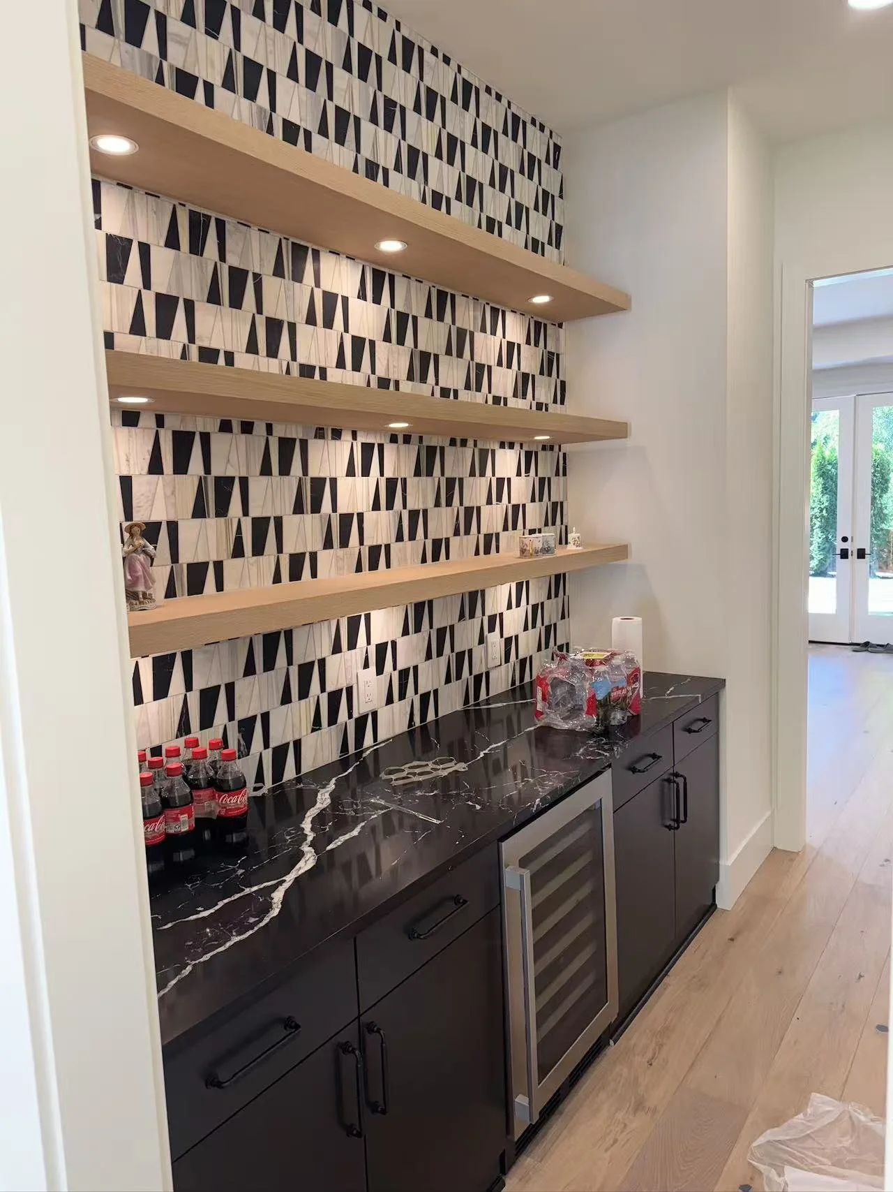 A modern kitchen bar area with a black marble countertop, black cabinets, and decorative shelves above with a geometric pattern wallpaper. There are bottles of Coca-Cola on the counter, and some items like a paper towel roll and a plastic wrapped pac