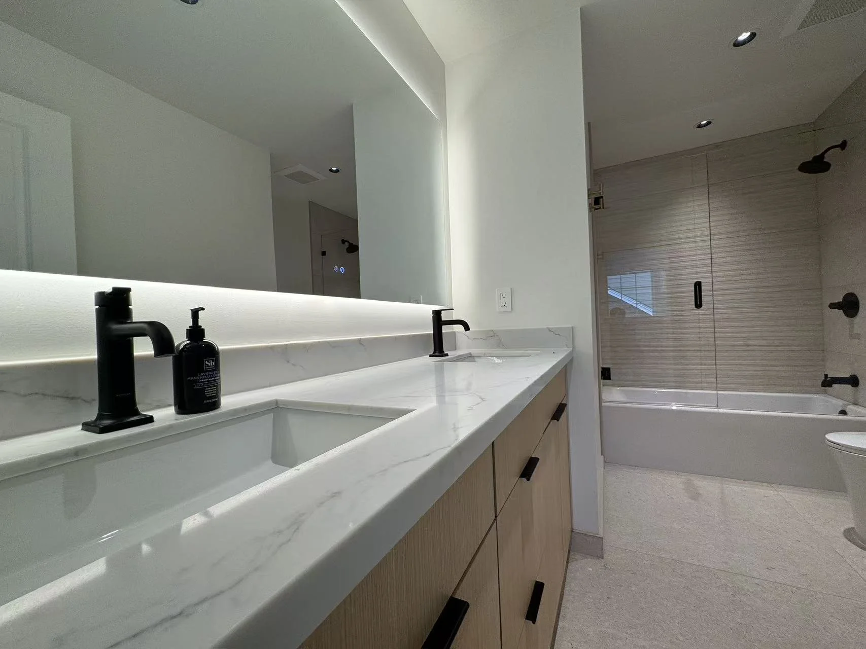 Modern bathroom with double vanity featuring black faucet and soap dispenser, large mirror, walk-in shower with glass door, beige textured wall, and white bathtub.