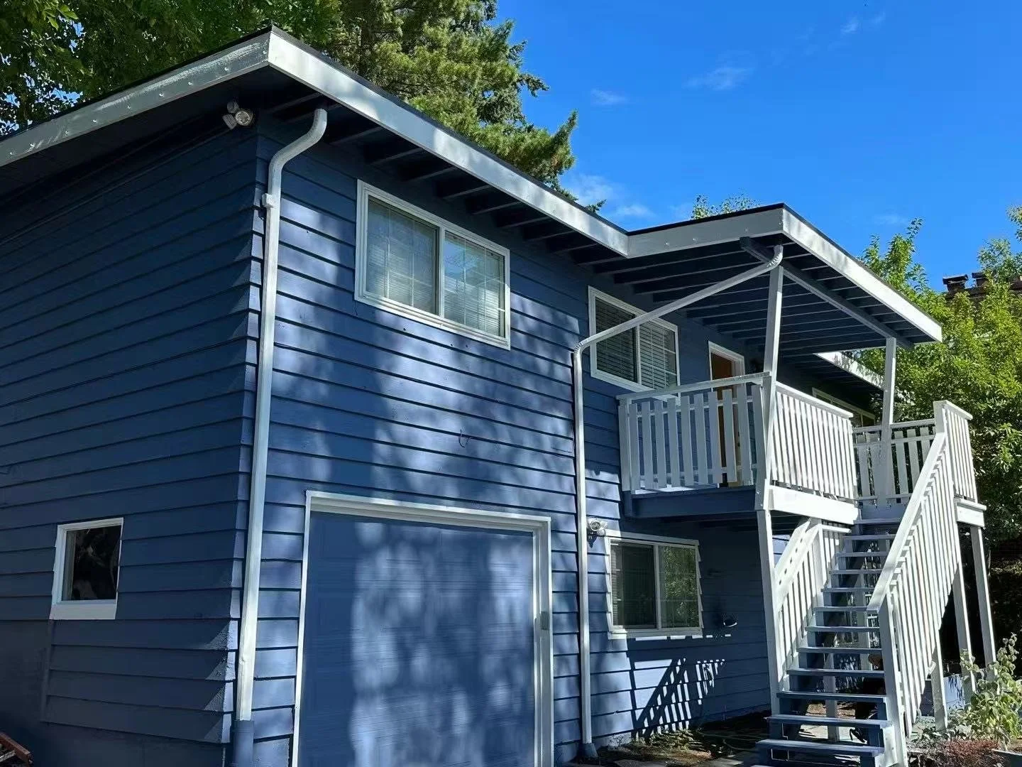 The back of a blue house with a white staircase and porch, wood siding, and two windows, under a clear blue sky with trees in the background.