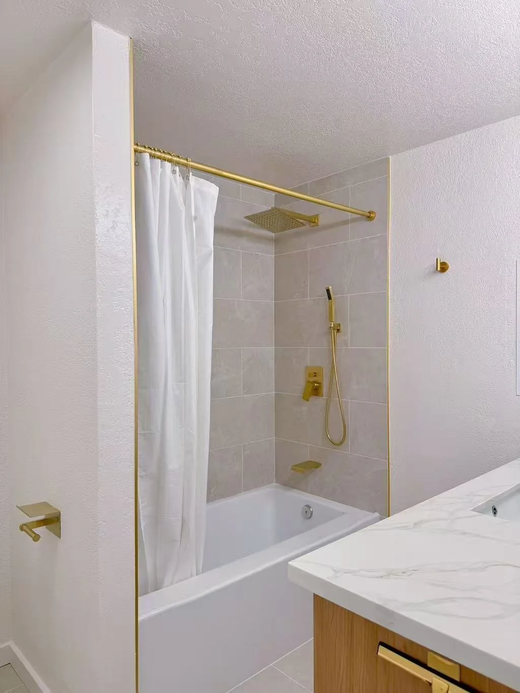 Modern bathroom with white bathtub, gold fixtures, shower with rainfall showerhead, and a white shower curtain, marble countertop, and neutral wall tiles.
