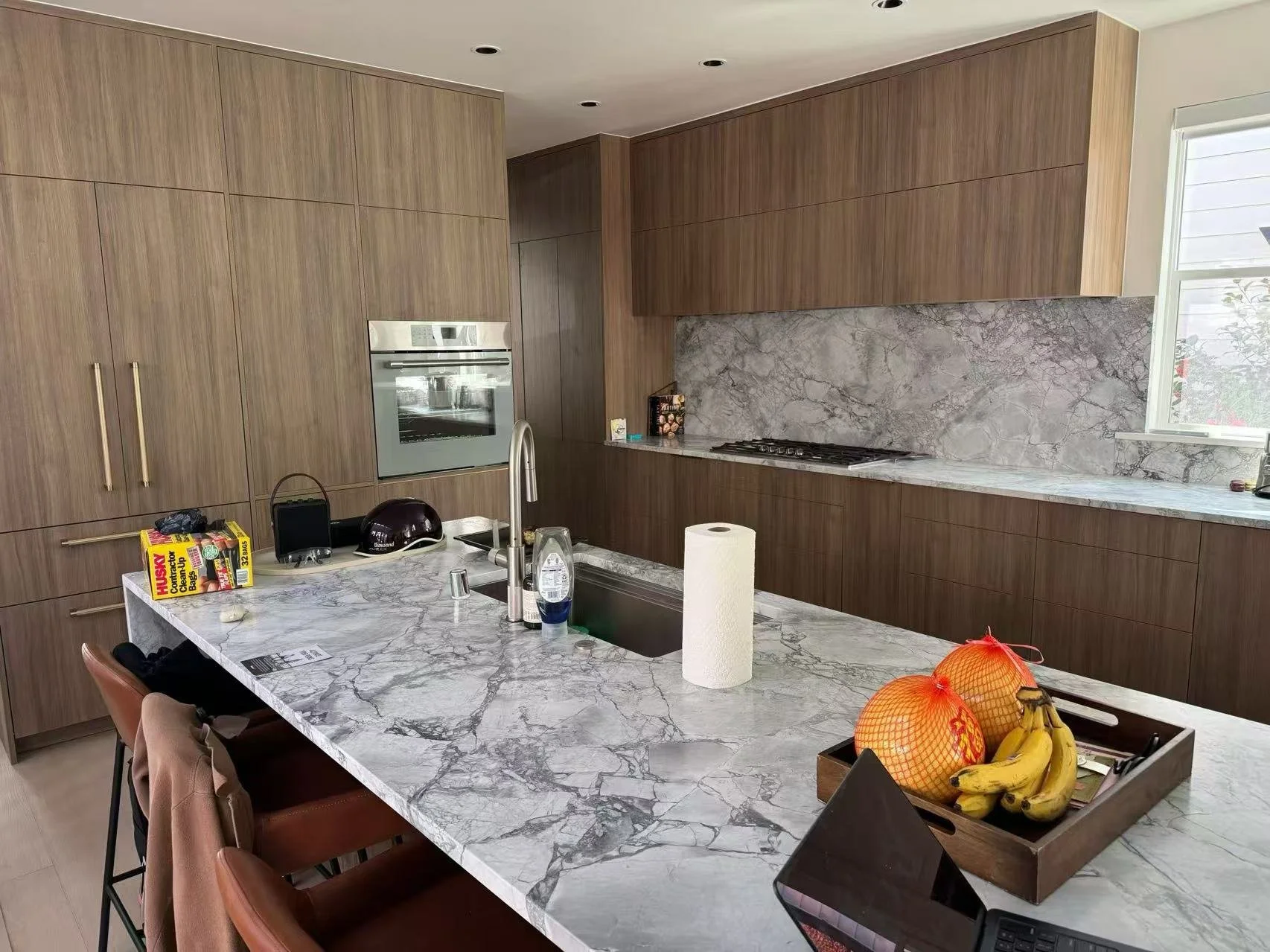 Modern kitchen with wooden cabinets, marble countertops, and a gray marble backsplash. There is a kitchen island with a sink, paper towel roll, and fruit basket containing bananas and oranges. On the island, there is also a laptop, a helmet, and a bo