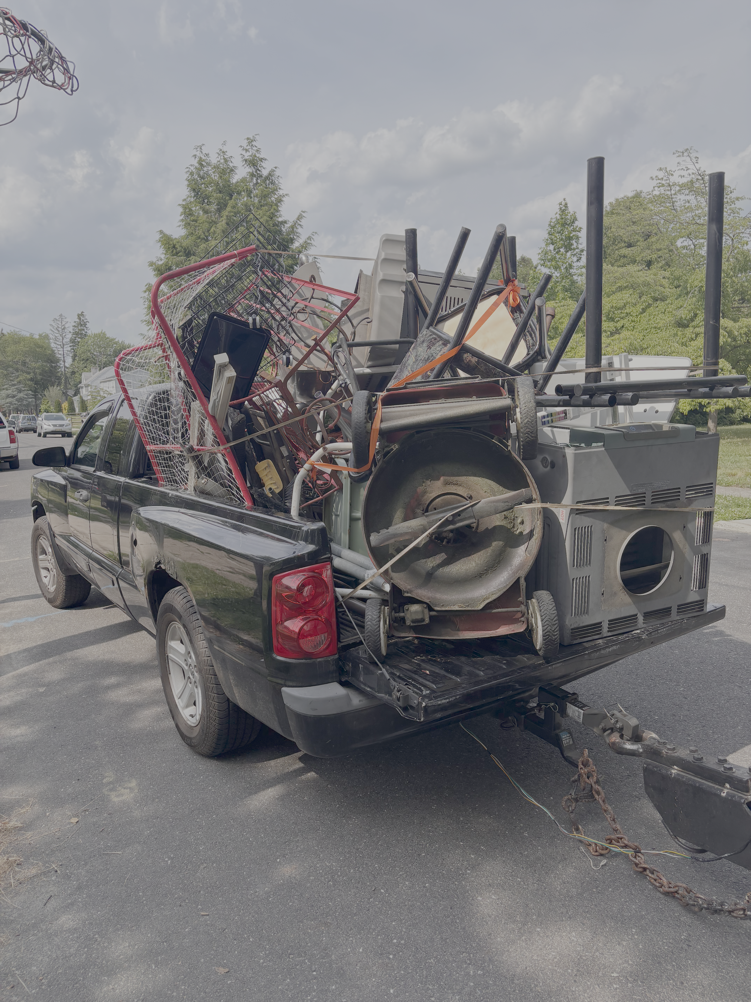 A black pickup truck with an open trailer hitch carrying a large pile of assorted shopping carts, some of which are damaged or bent, parked on an asphalt lot with trees and a residential neighborhood in the background.