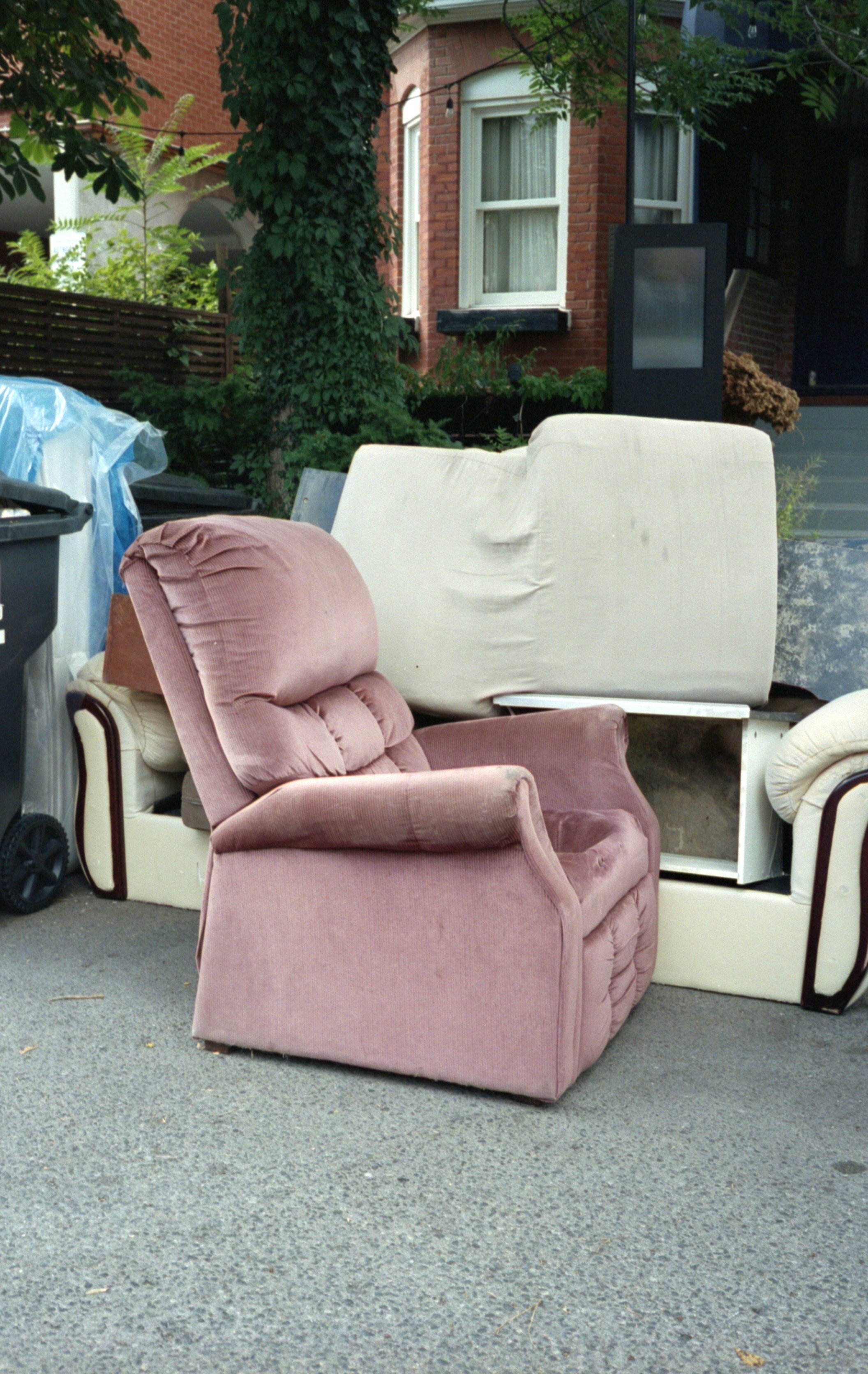 Various used armchairs and sofas stacked outdoors on a street against a backdrop of a brick house and green trees, with a trash bin and miscellaneous items nearby.