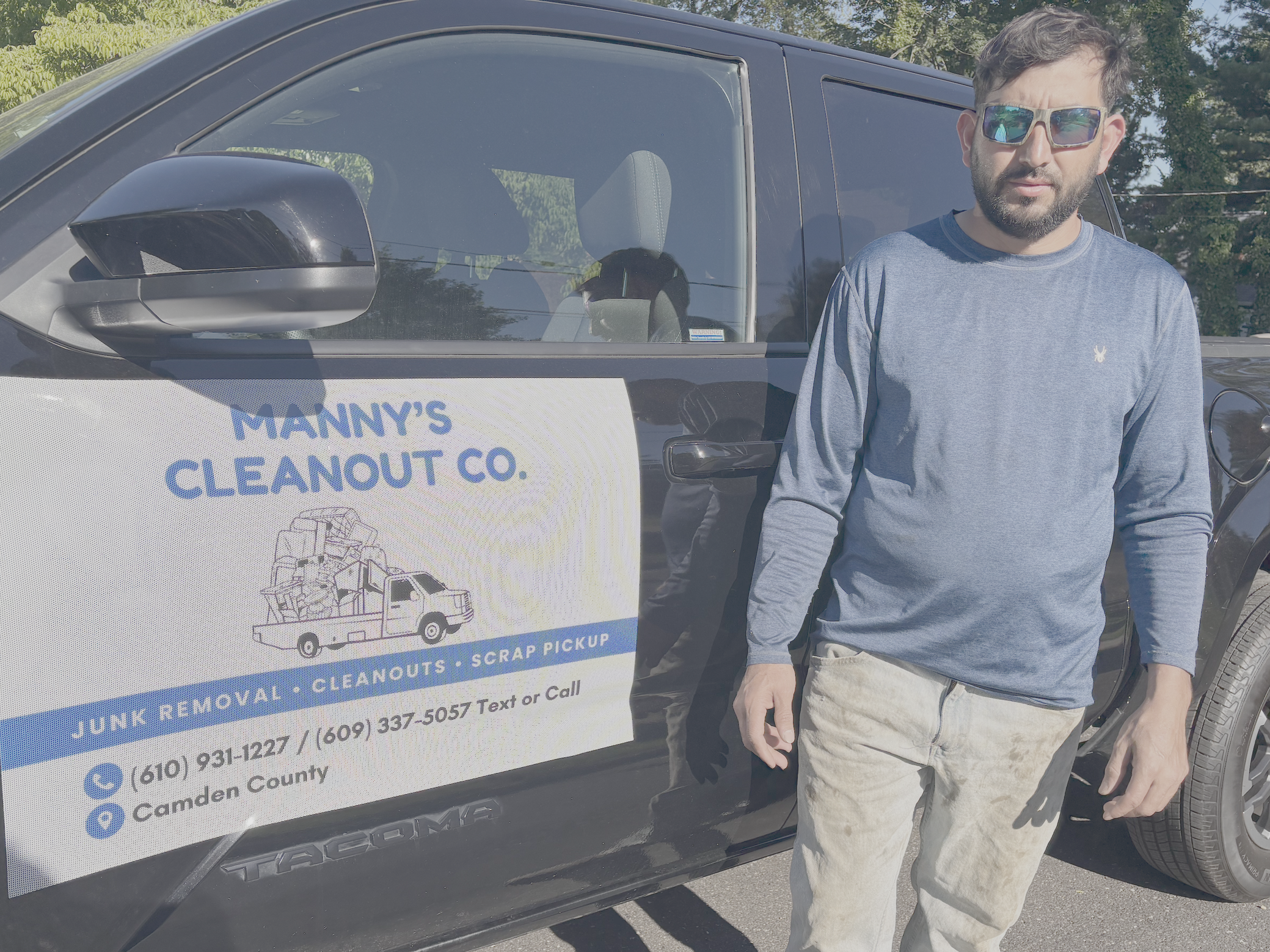 A man standing next to a black van with a sign for Manny's Cleanout Co. shows phone number and services such as junk removal, cleanouts, and scrap pickup. The man wears sunglasses, a gray long-sleeve shirt, and light-colored pants, outdoors with green trees and sky in the background.