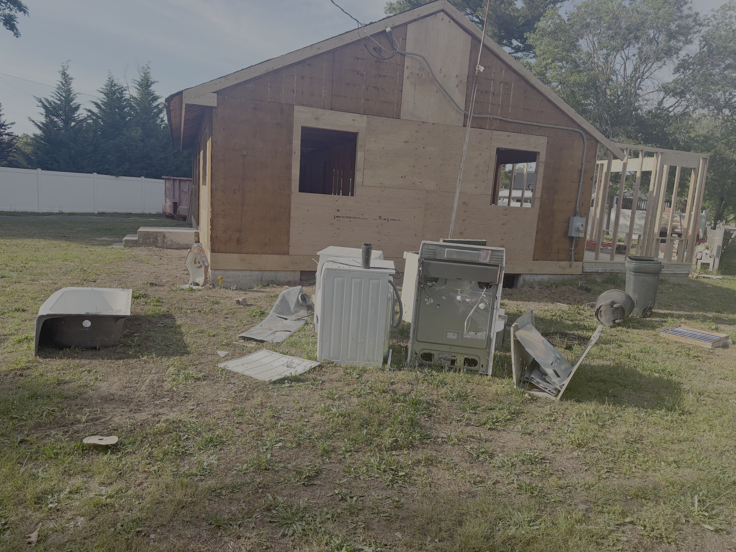 A house under construction with unfinished wooden walls and a sloped roof. Construction debris and tools are scattered around the yard, and a white fence surrounds the area.