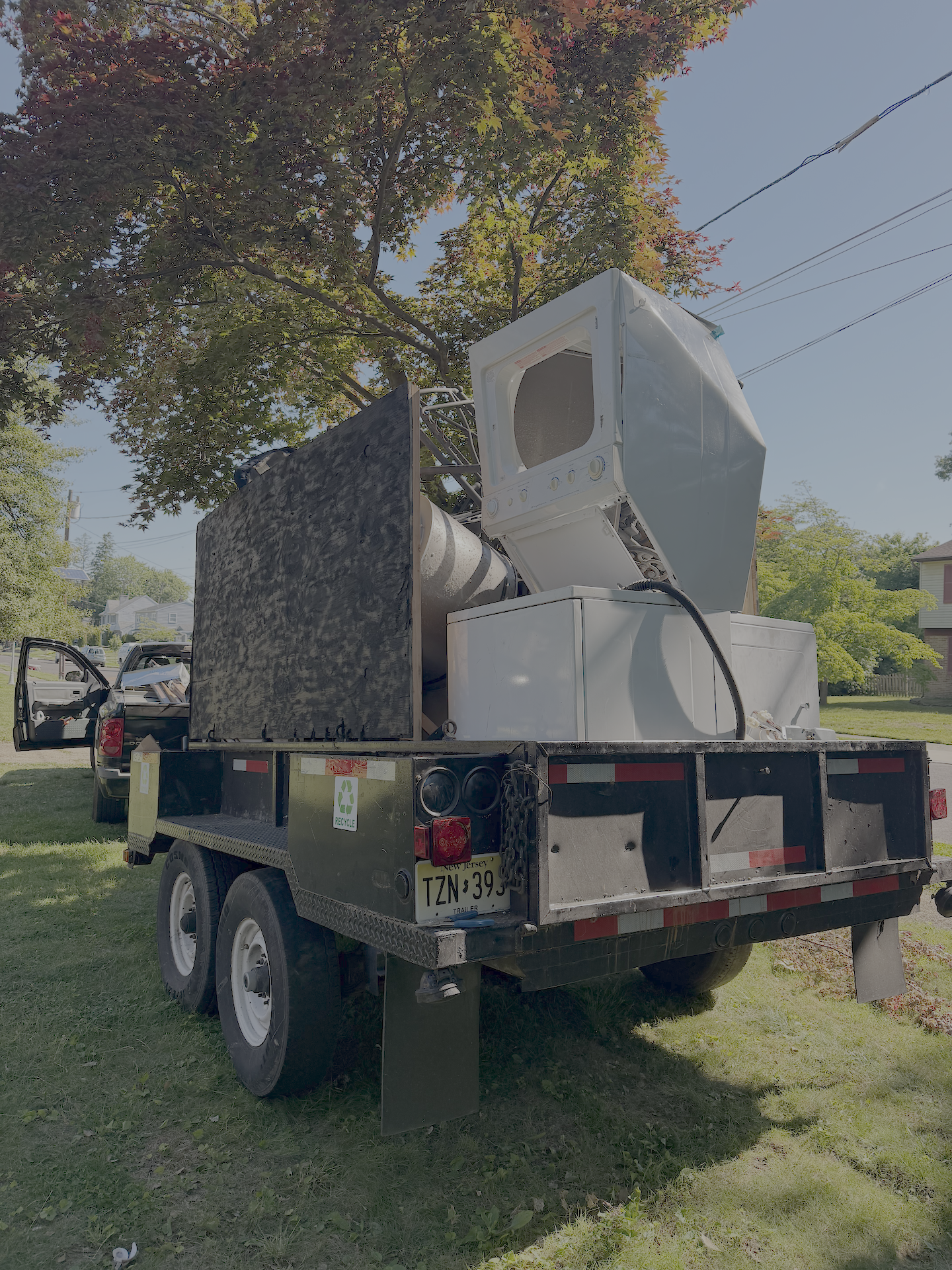 A flatbed truck parked on a grassy area with a washing machine and other appliances loaded on it. There are trees and residential houses in the background.