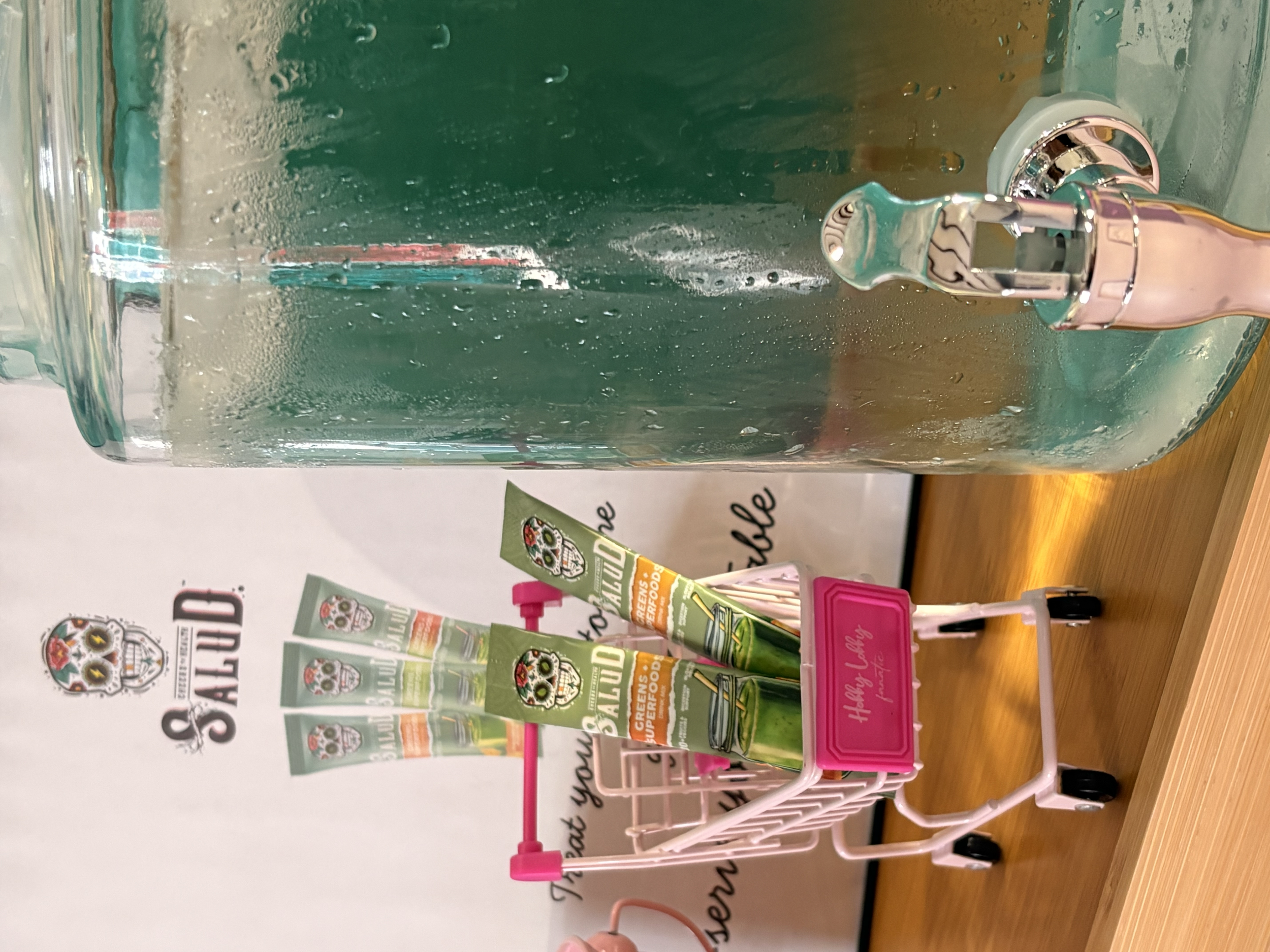 A display of colorful tube-shaped products with green and orange labels, possibly containing food or supplements, on a small pink rack next to a glass container with water and condensation on its surface. Behind the display is a partially visible sig