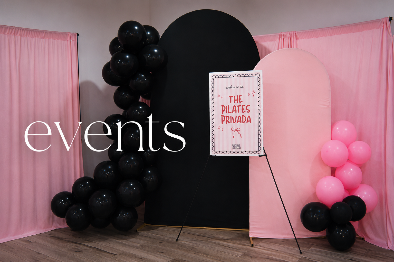 Decorative event backdrop with pink curtains, black and pink balloons, a sign for 'The Pilates Privada' gym, and a black and pink color scheme.