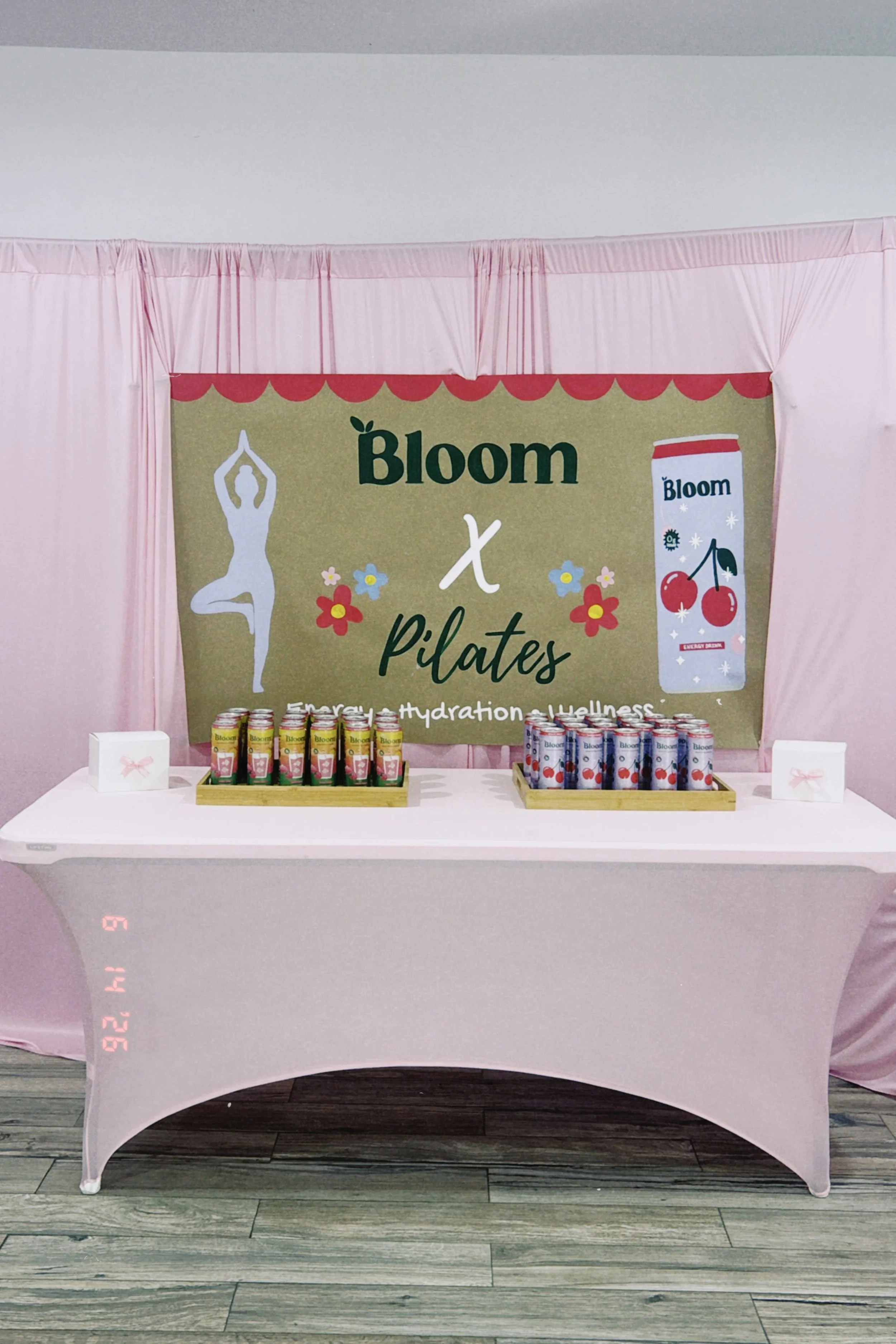 Table covered with a pink tablecloth displaying cans of Bloom beverage, with a backdrop labeled 'Bloom x Pilates' and a yoga figure illustration, in an indoor setting.