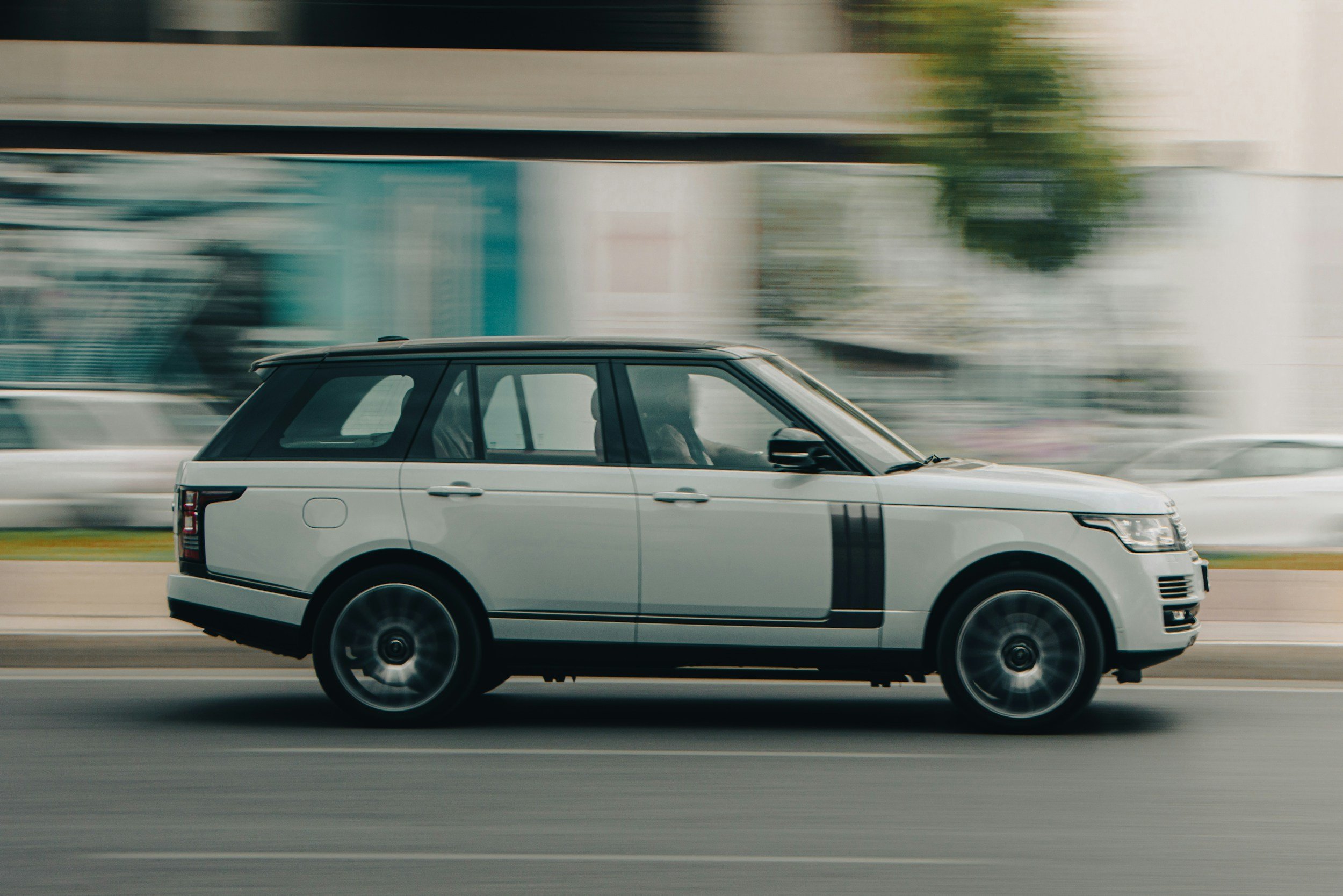 A white SUV driving on a road with a blurred background indicating motion, trees, and urban structures.