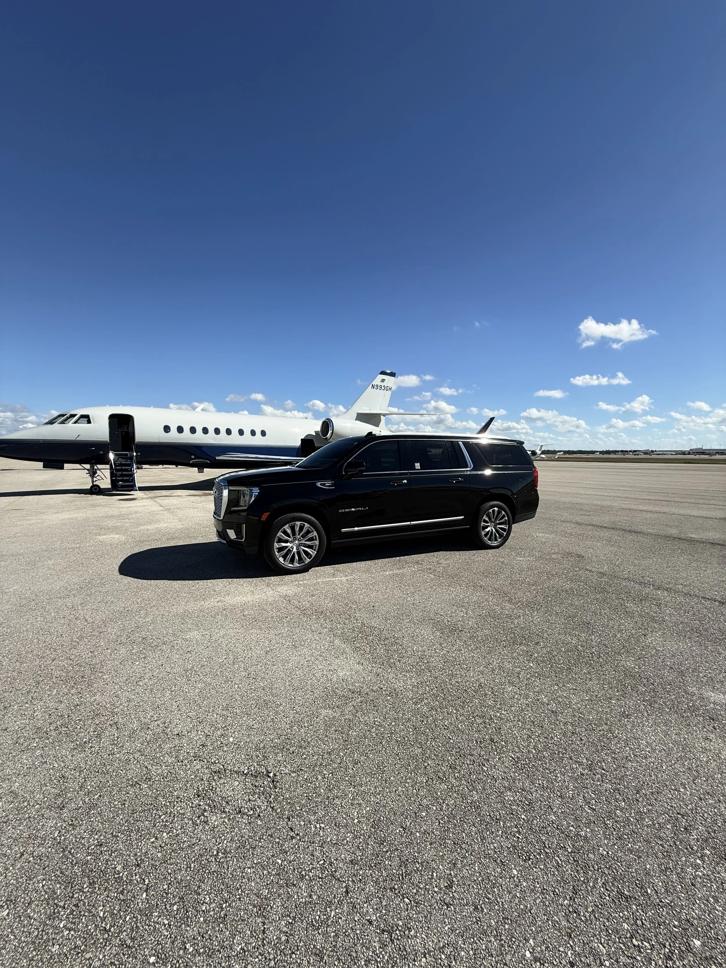 A private jet parked on an airport tarmac with a black SUV nearby under a blue sky with some clouds.