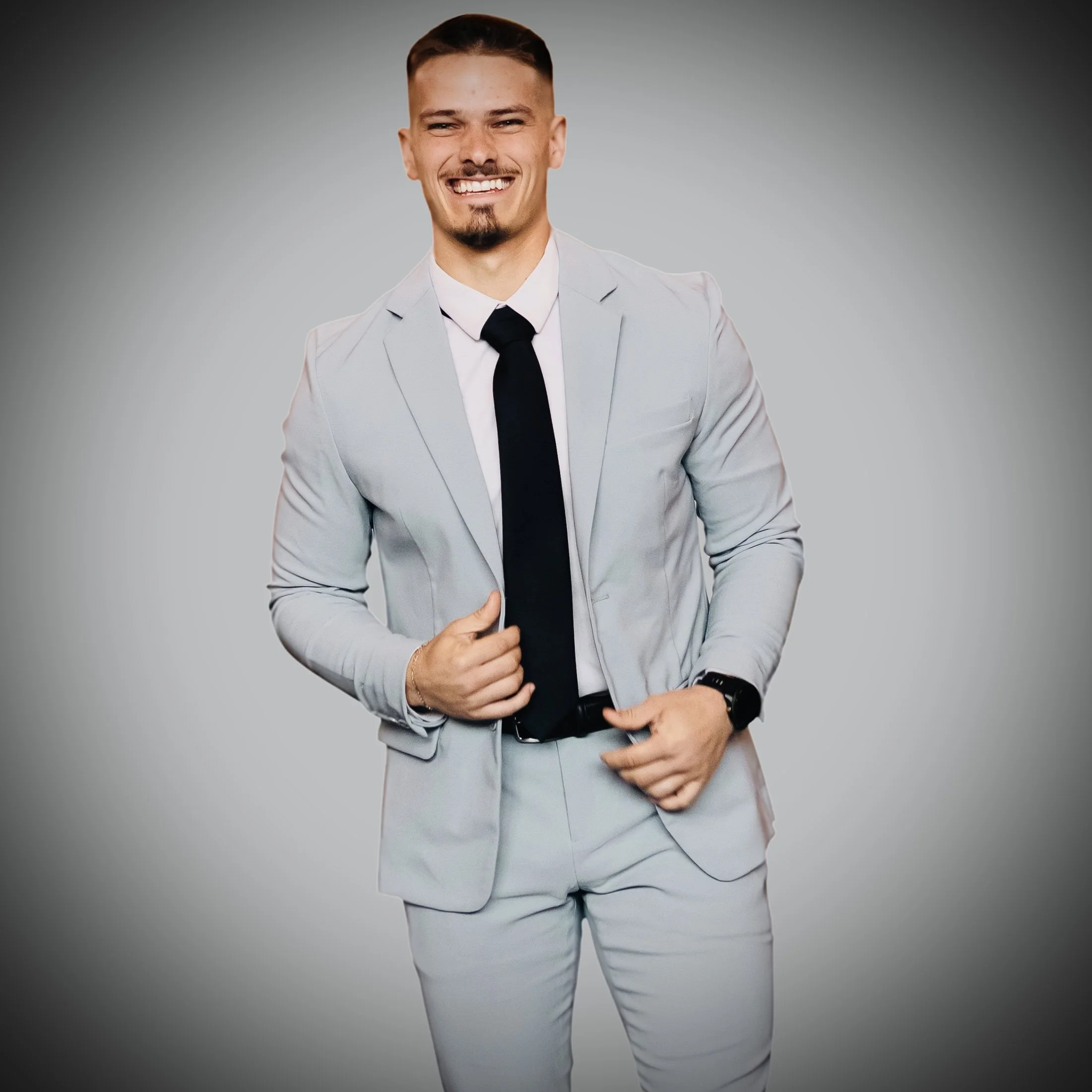 A man in a light gray suit with a white shirt and black tie, smiling and posing with one hand on his stomach against a plain gray background.