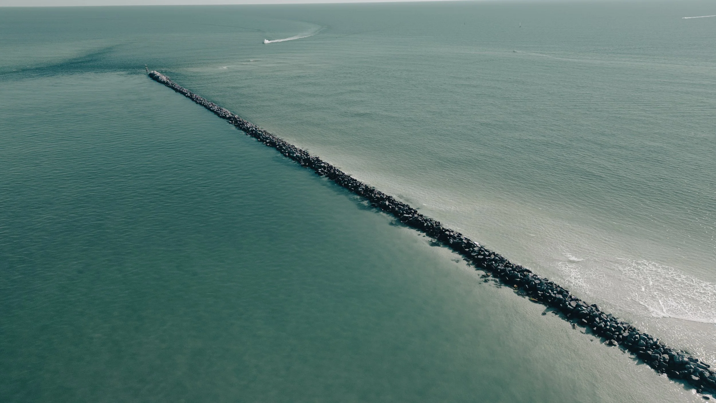 An aerial view of a long rock jetty extending into the ocean with boats visible in the distance, creating wakes on the water.