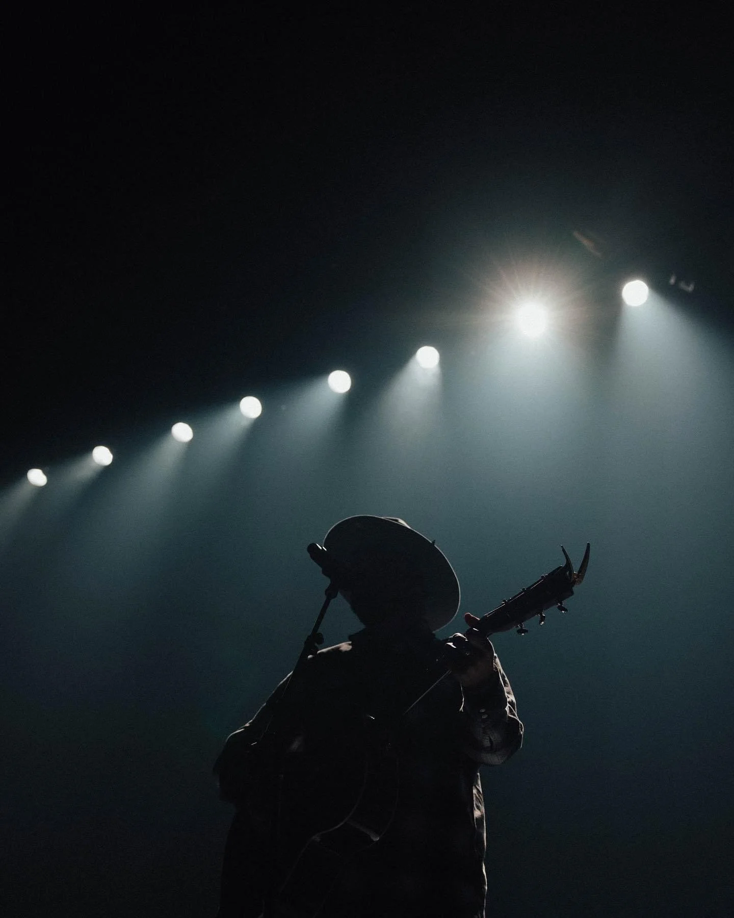 Silhouette of a person playing guitar on stage under bright white spotlights in a dark setting.