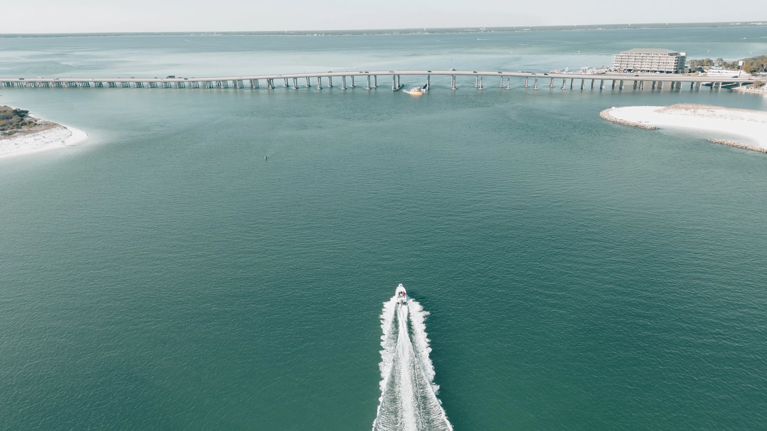 A boat speeds through calm waters near a bridge with multiple supports, with a sandy beach and hotel in the background under a clear sky.