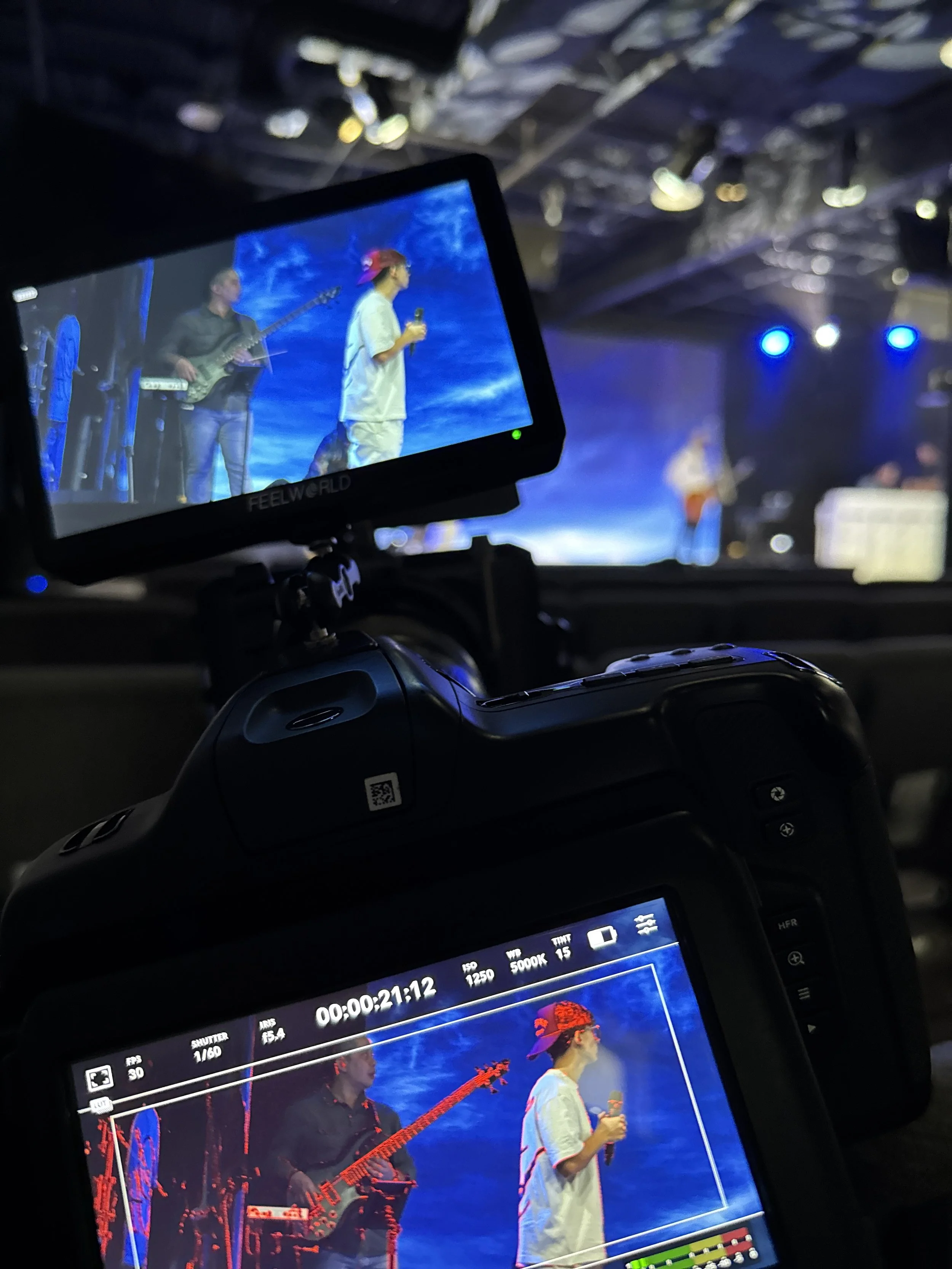 A concert scene is visible through a camera's screen showing a musician with a red cap and white shirt holding a microphone, and another musician in black playing an electric guitar. The background is blue with stage lights. The camera is mounted inside a vehicle, with part of a dashboard display visible.