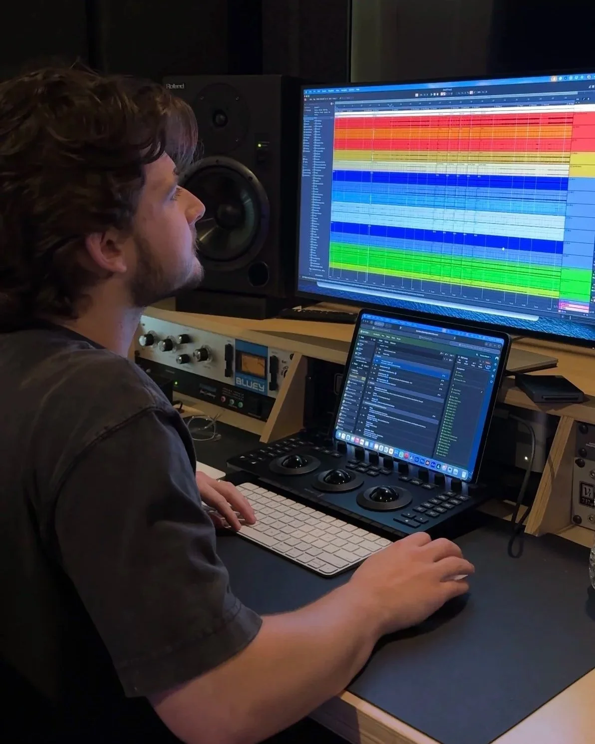 Man working on music production in a studio, with a large screen displaying a colorful digital audio workstation and a smaller monitor showing email or project files.