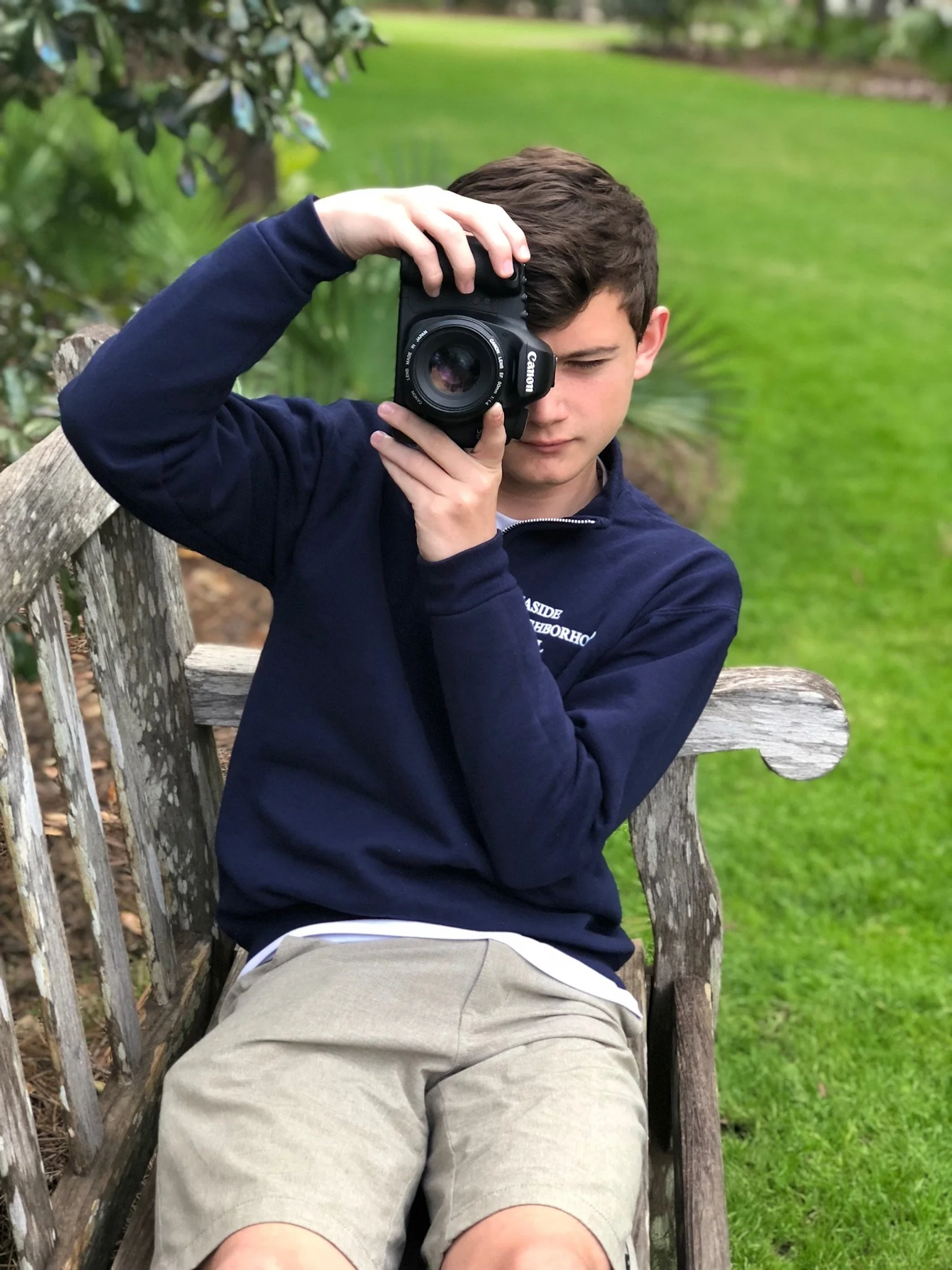 A young man sitting on a wooden bench outdoors, holding a Canon camera to his face, looking through the viewfinder, with green grass and bushes in the background.