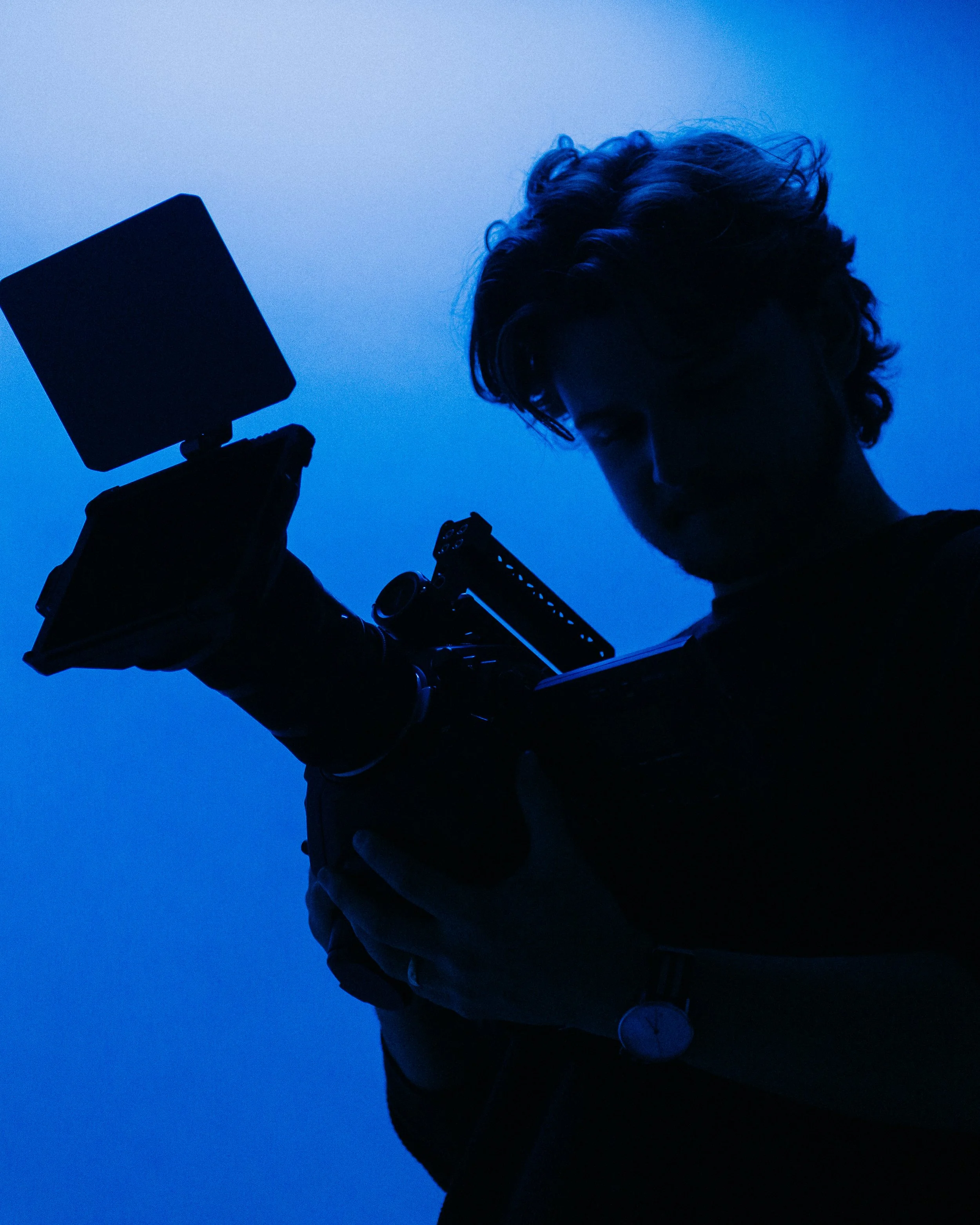 Silhouetted person with curly hair holding a camera and a smartphone against a blue evening sky.
