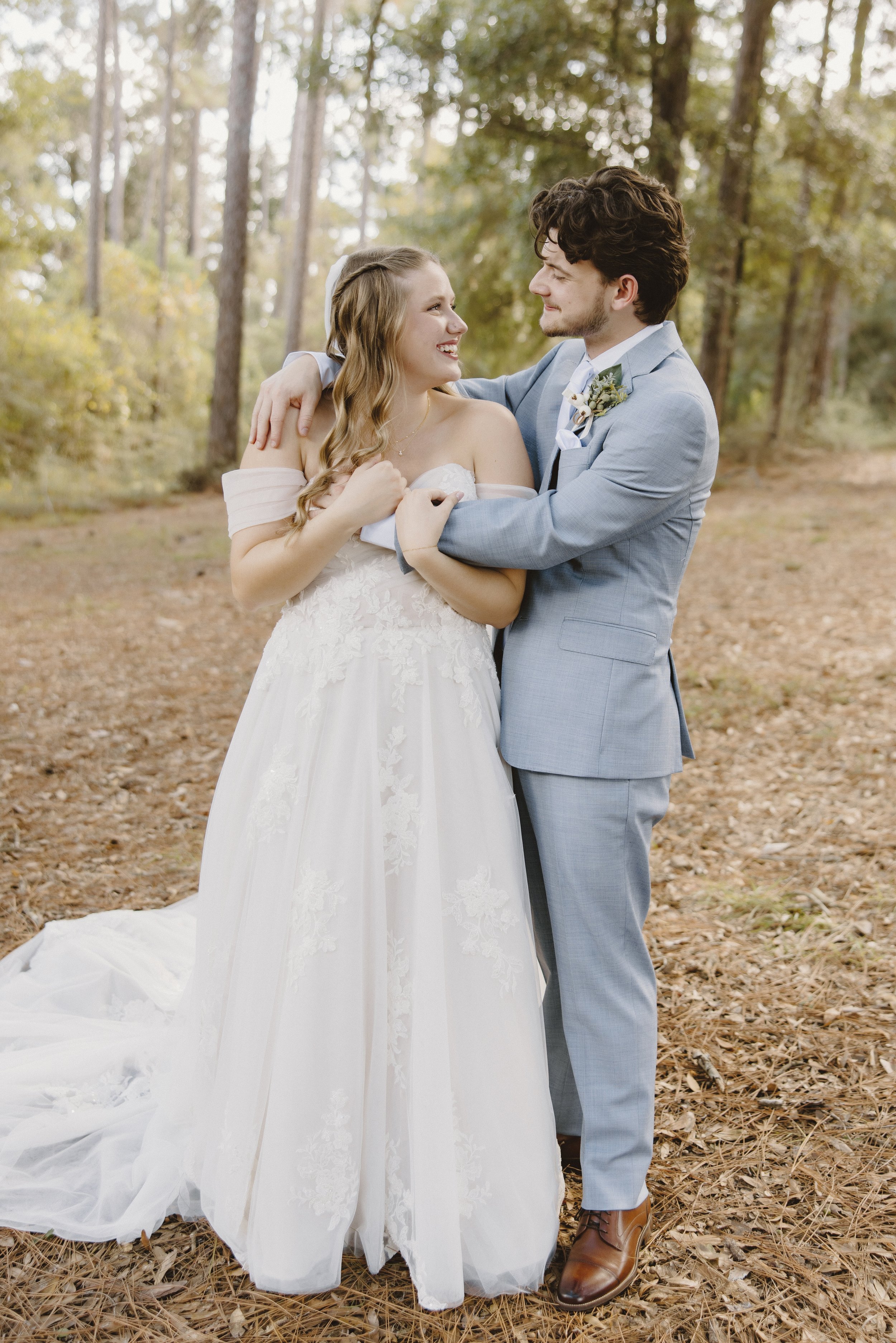 A bride and groom share a dance outdoors in a forested area with trees and fallen leaves, smiling at each other.