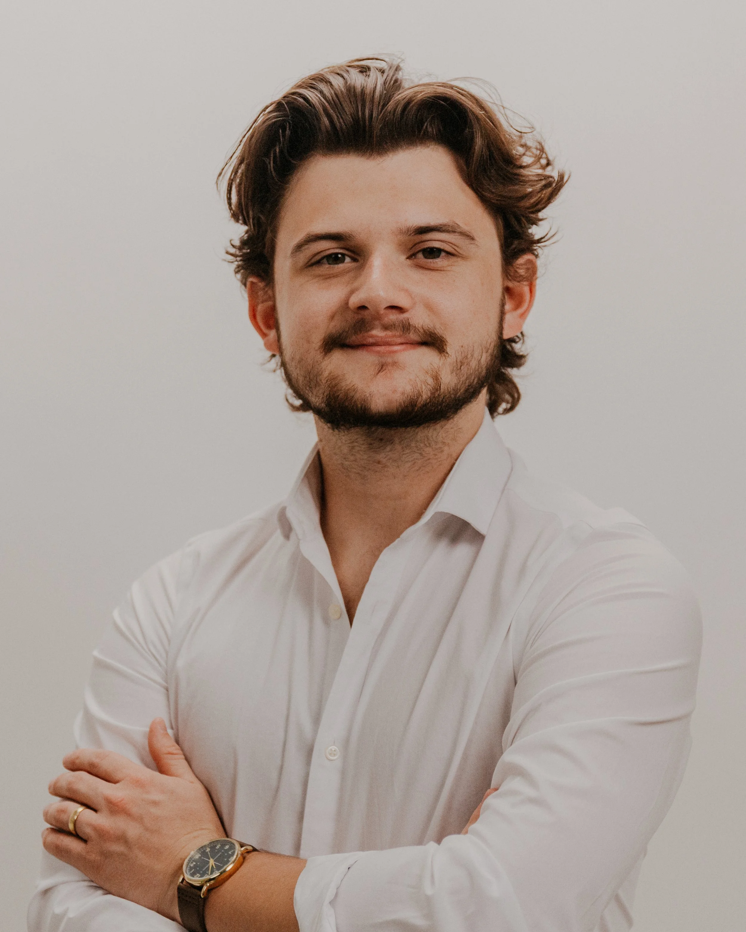 Close-up of a young man with wavy brown hair, dressed in a white shirt, smiling gently, with arms crossed, against a plain white background.