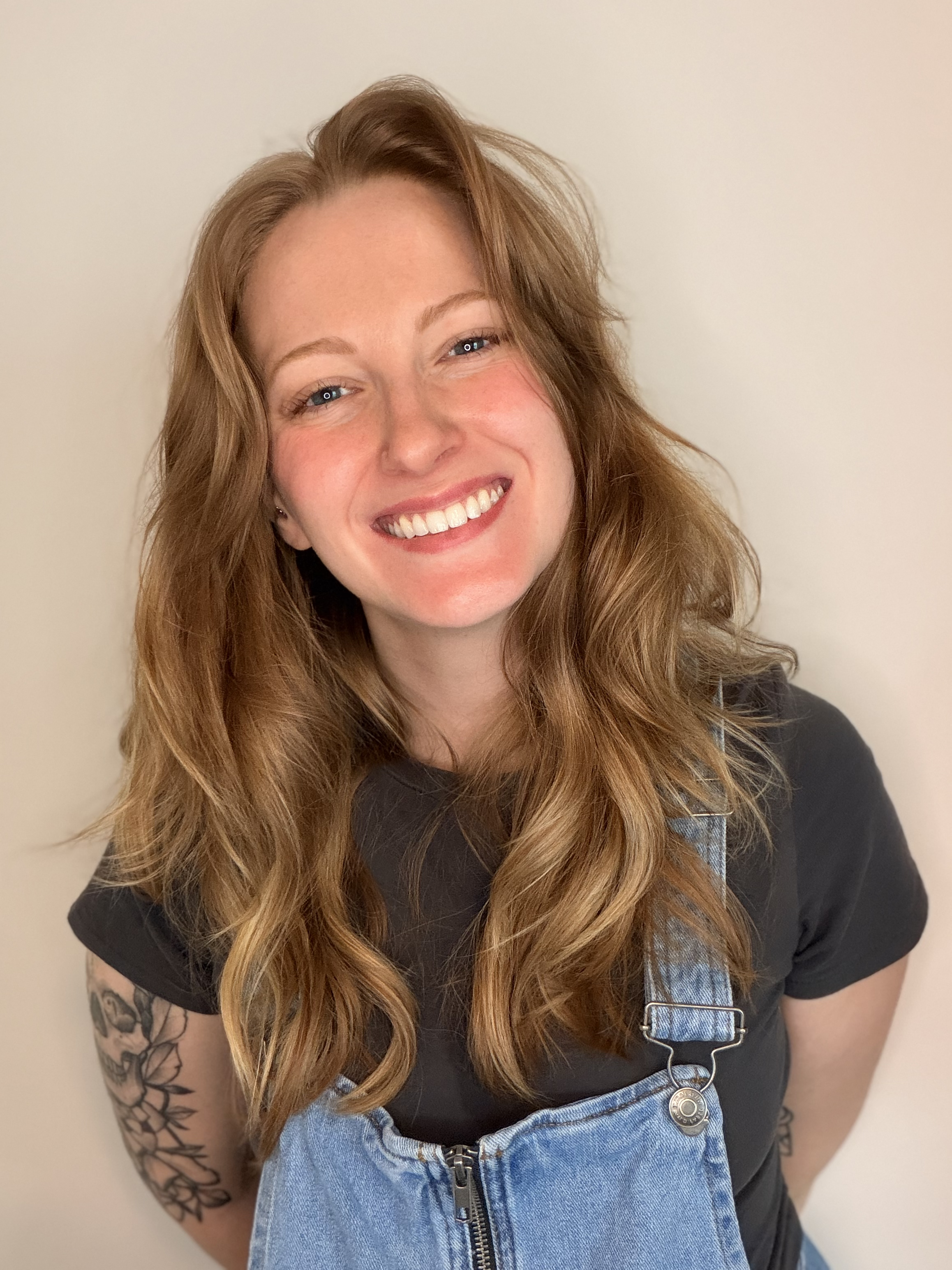 A young woman with long wavy red hair smiling, wearing a black t-shirt and blue denim overalls, with floral tattoos on her arms, standing against a plain white background.