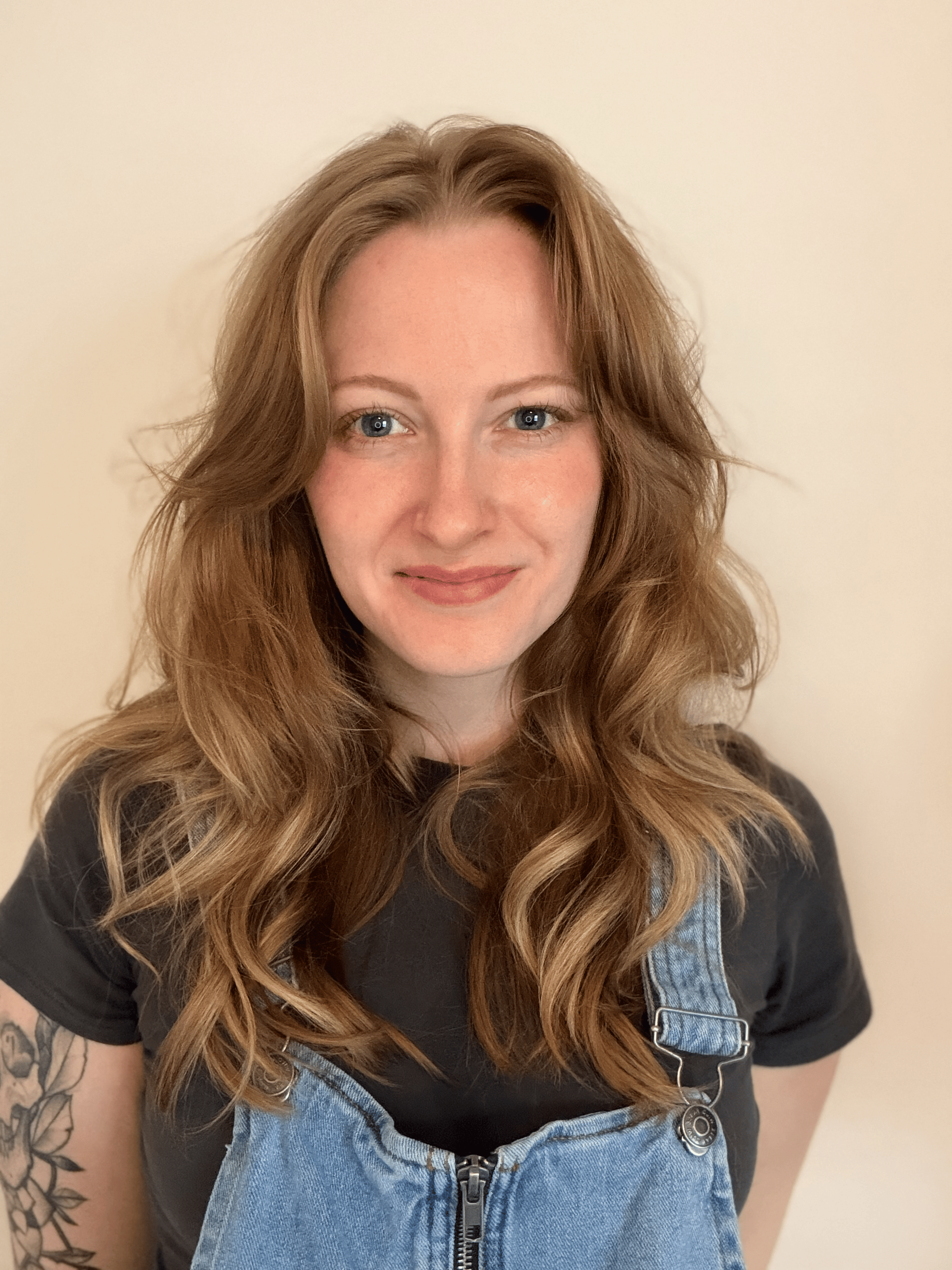 Close-up of a woman with long wavy auburn hair, wearing a black t-shirt and denim overalls, with a floral tattoo on her left arm, smiling against a plain wall background.