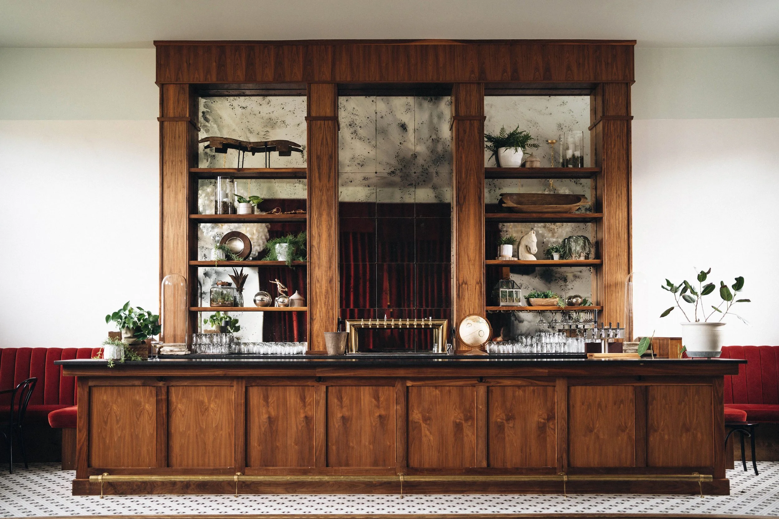 A wooden bar with glassware and plants, red chairs, and a white tiled floor in a restaurant or lounge setting.