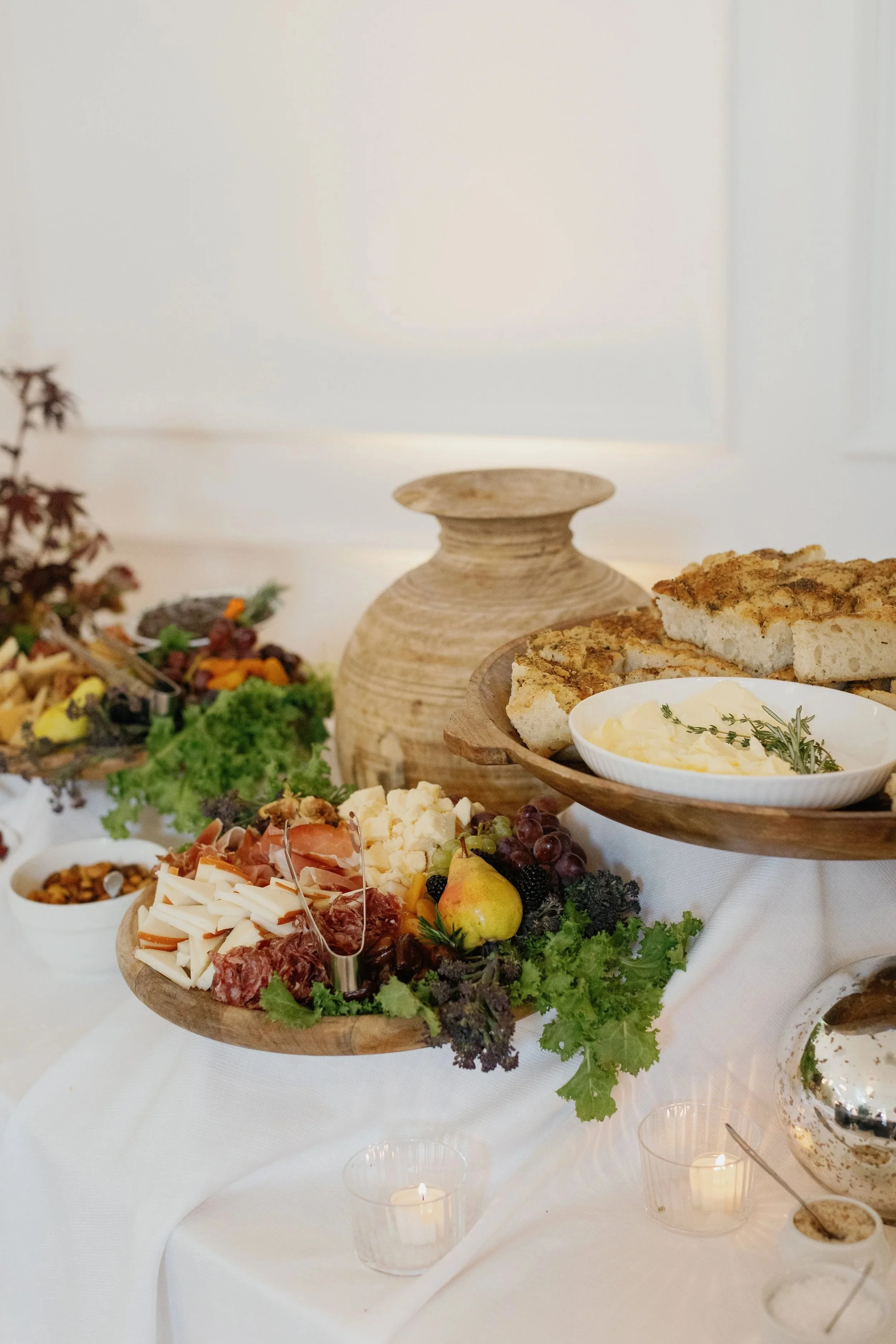 A buffet table with cheese, meats, fruits, vegetables, bread, and dips, decorated with greenery and lit by candles.