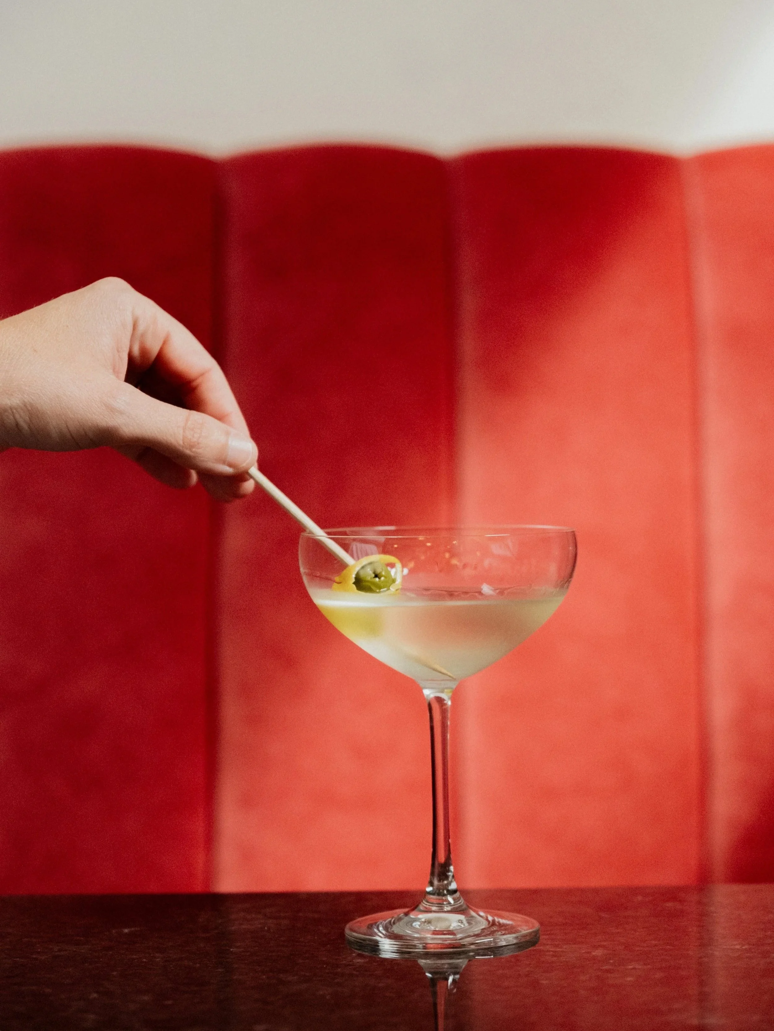 A hand holding a cocktail pick garnished with an olive, positioned above a coupe glass filled with a clear beverage, with a red and white background.