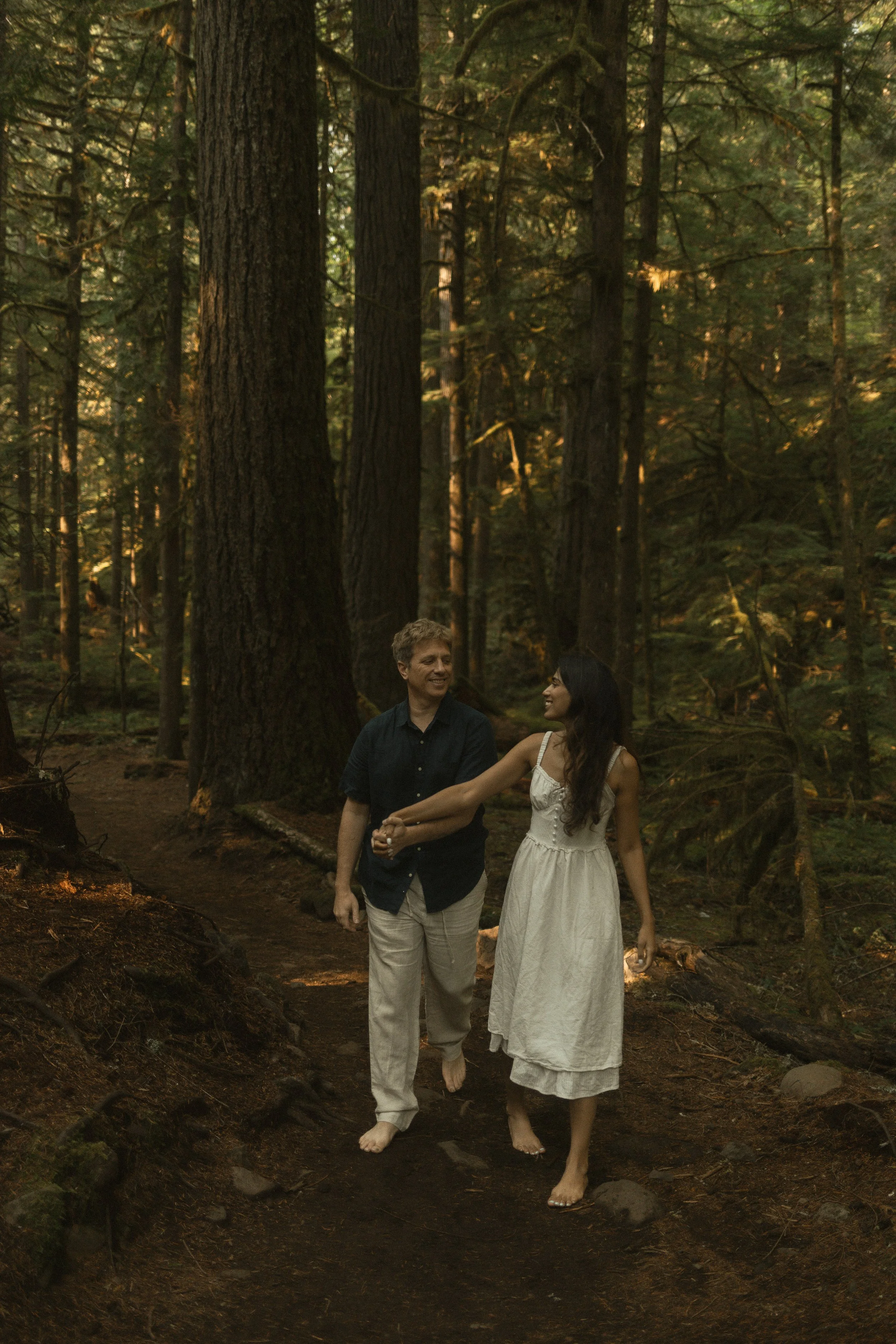 Two people, a man and a woman, walking barefoot in a forest. The man is wearing a dark shirt and light-colored pants, and the woman is dressed in a white dress. They are holding hands and smiling at each other. Tall trees surround them, and sunlight filters through the leaves.
