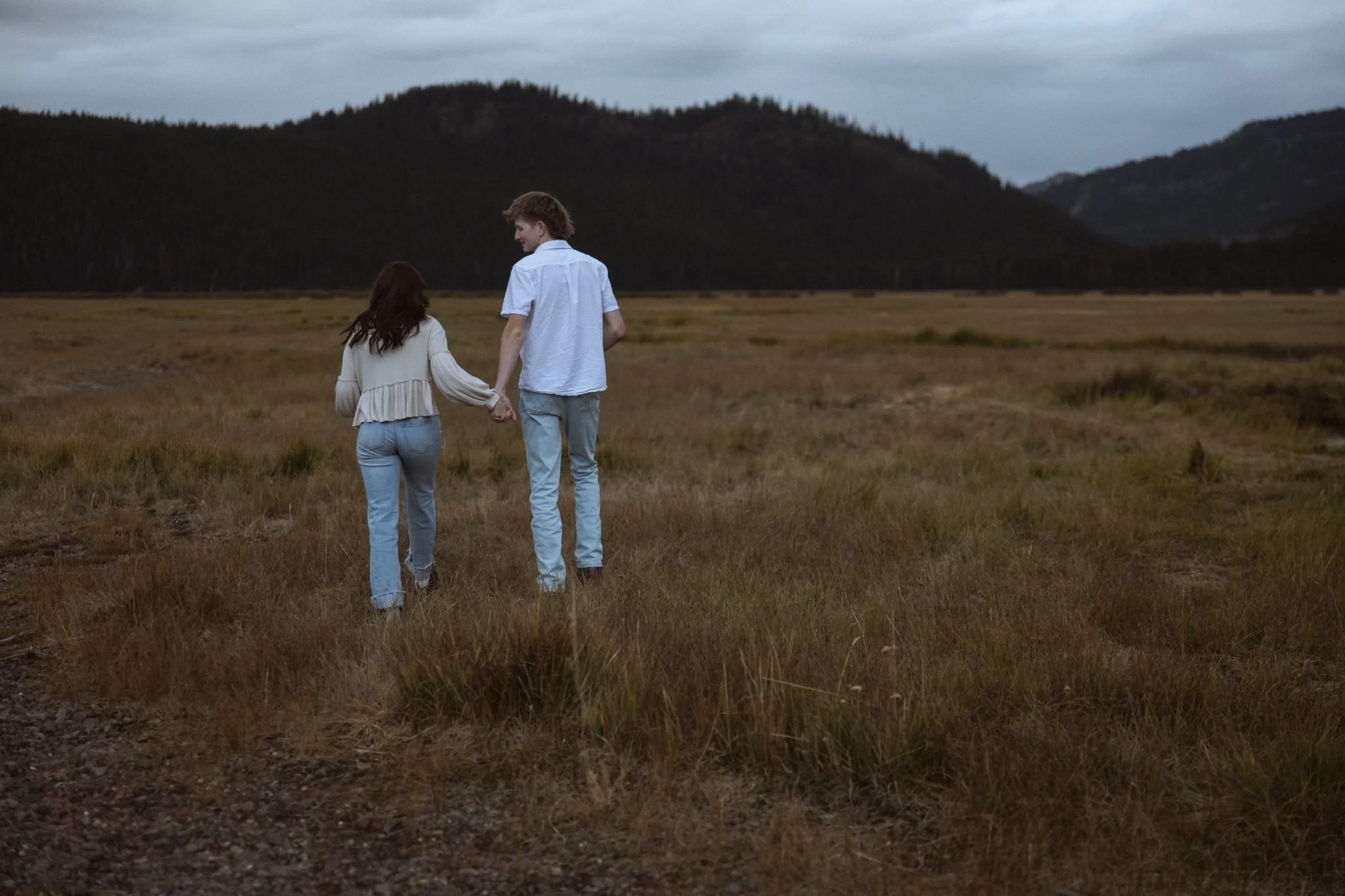 A young man and woman walk hand in hand across a grassy field with mountains in the background under a cloudy sky.