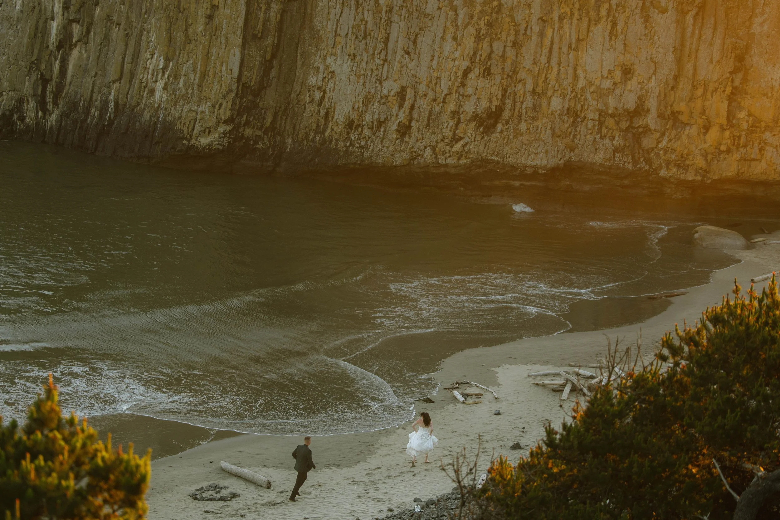 A couple dressed in wedding attire walking on a sandy beach near rocky cliffs and ocean waves, with some greenery in the foreground.