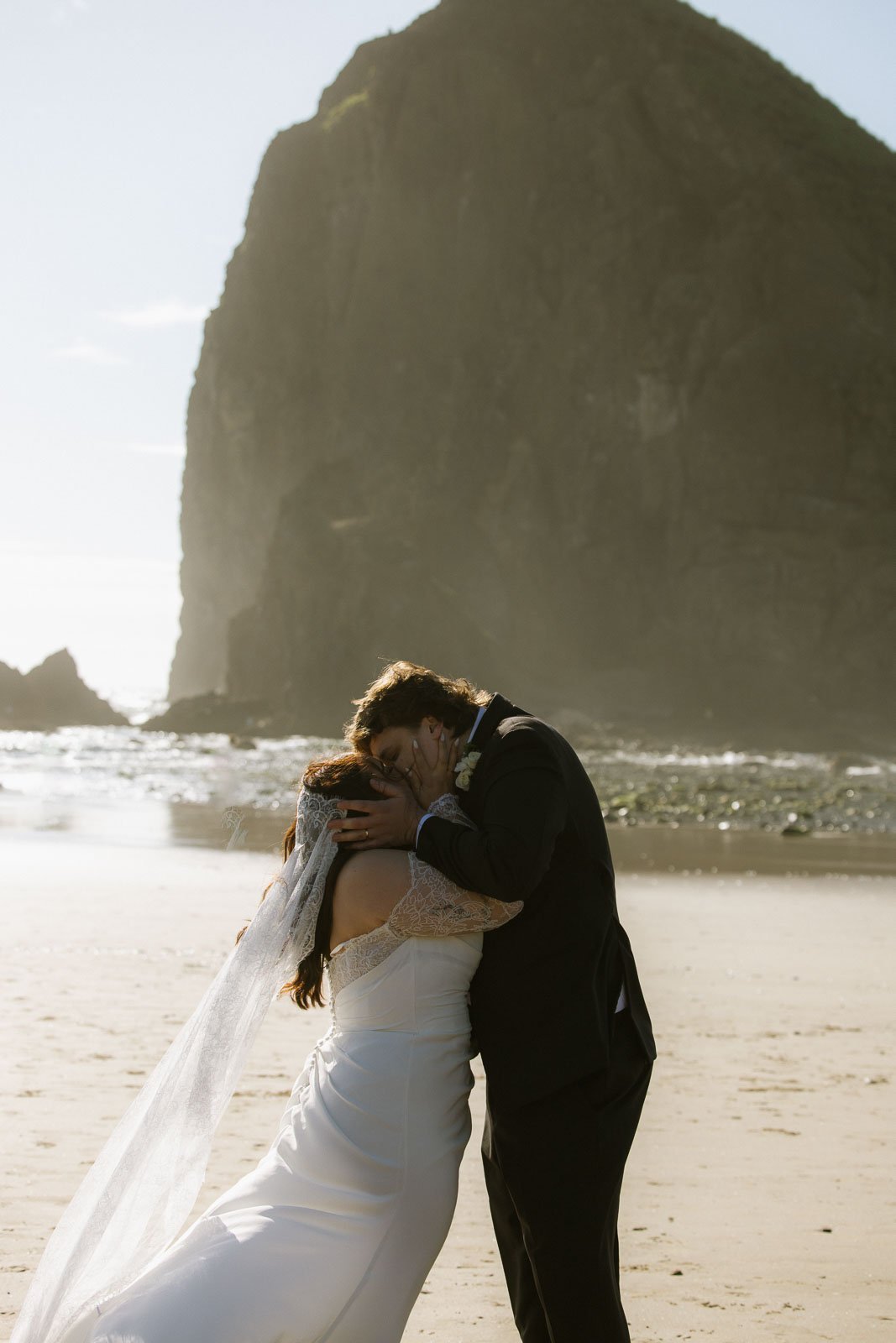 A bride and groom sharing a kiss and embrace on a beach with a large rock formation in the background during sunset.