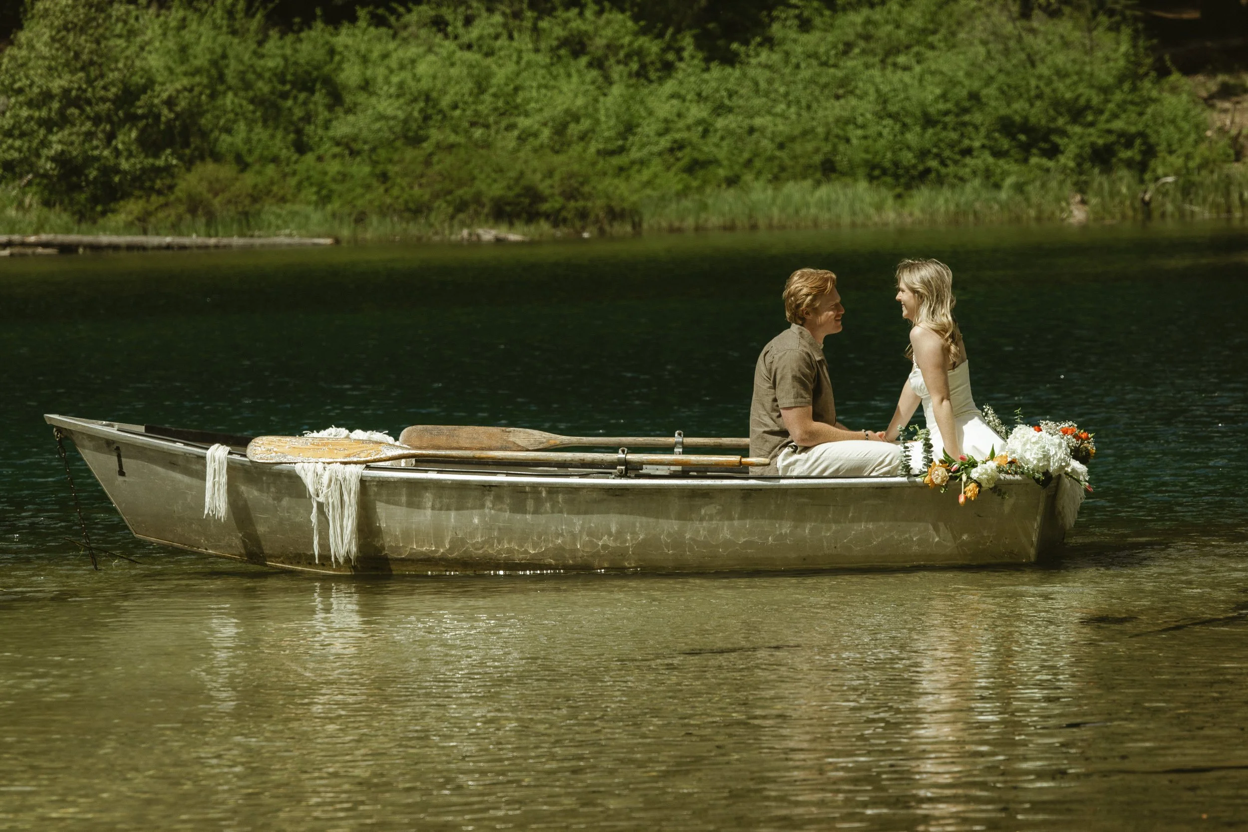 A couple sitting in a small boat on a calm lake, facing each other and smiling, with green trees in the background.