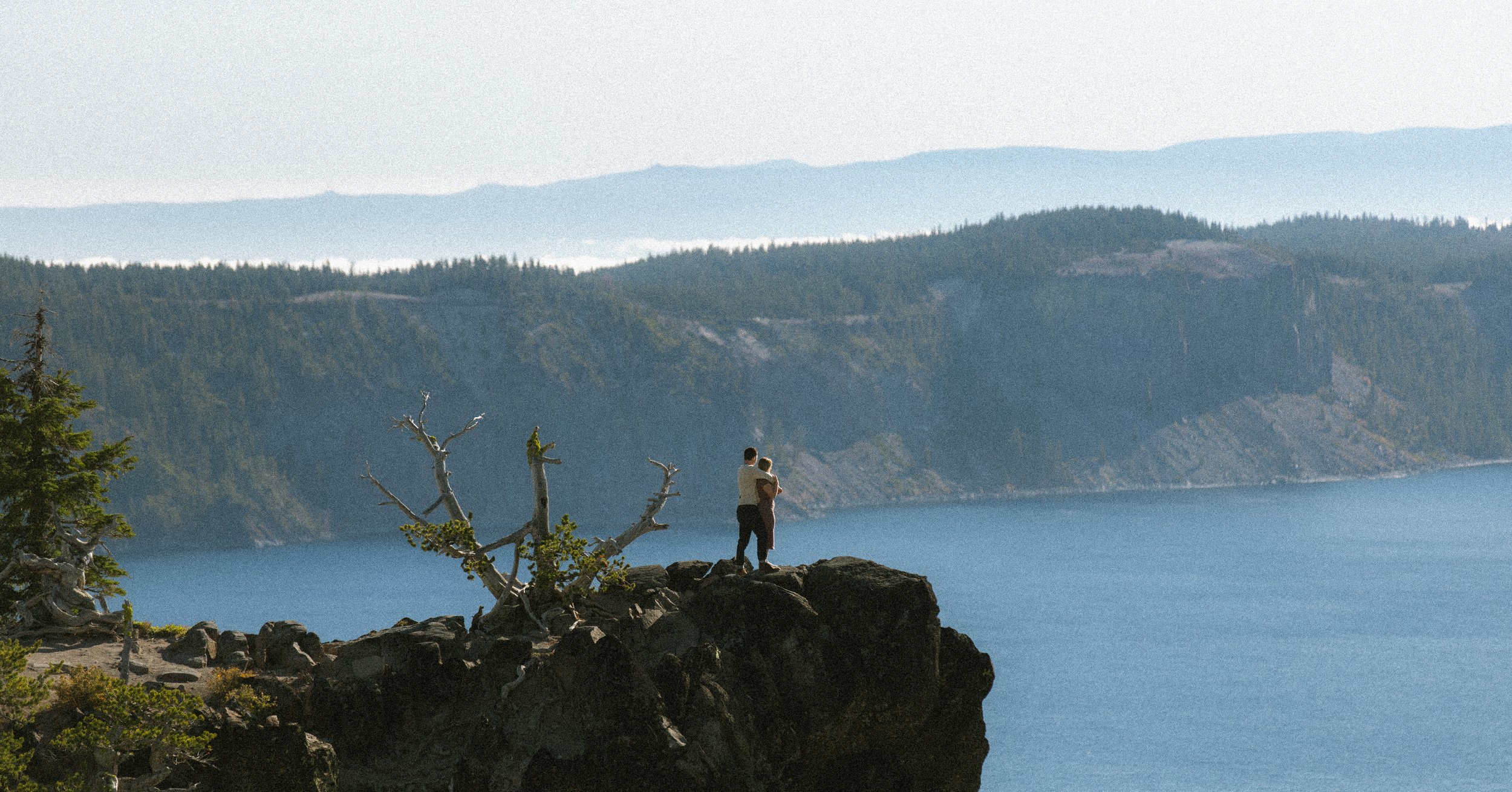 Two people standing on rocks by the water near a mountain range, with trees and a cloudy sky in the background.