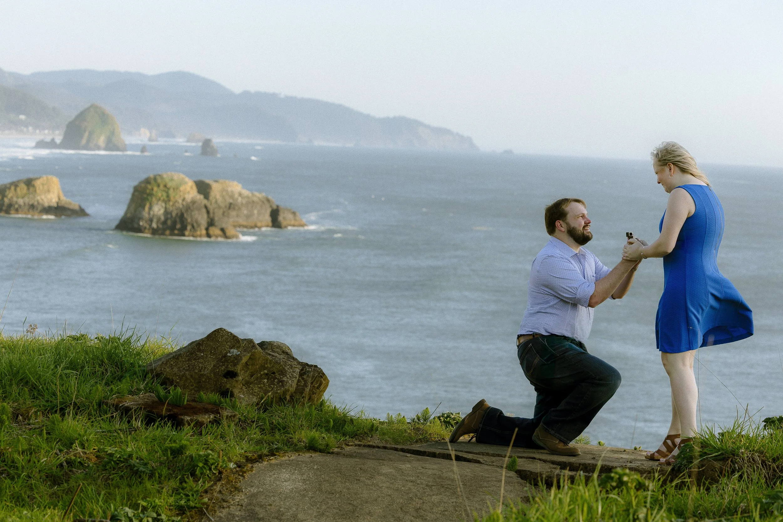 A man kneeling on one knee proposing marriage to a woman in a blue dress at a scenic coastal overlook with rocky islands and ocean in the background.