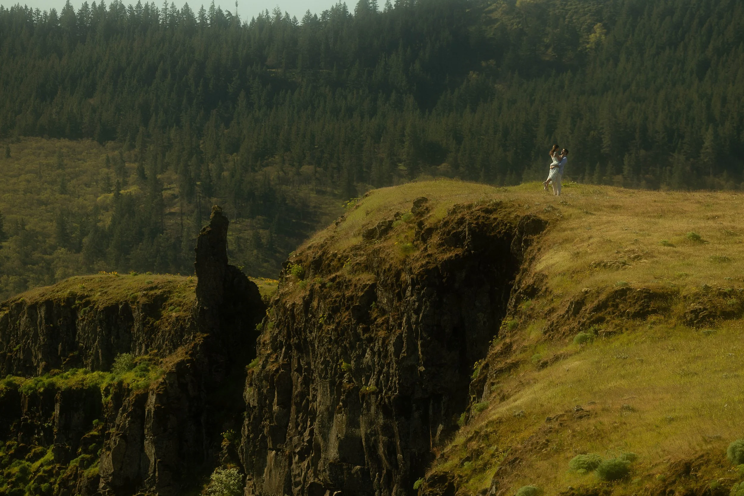 A couple dancing on a grassy cliff edge overlooking a forested canyon with steep rock walls.