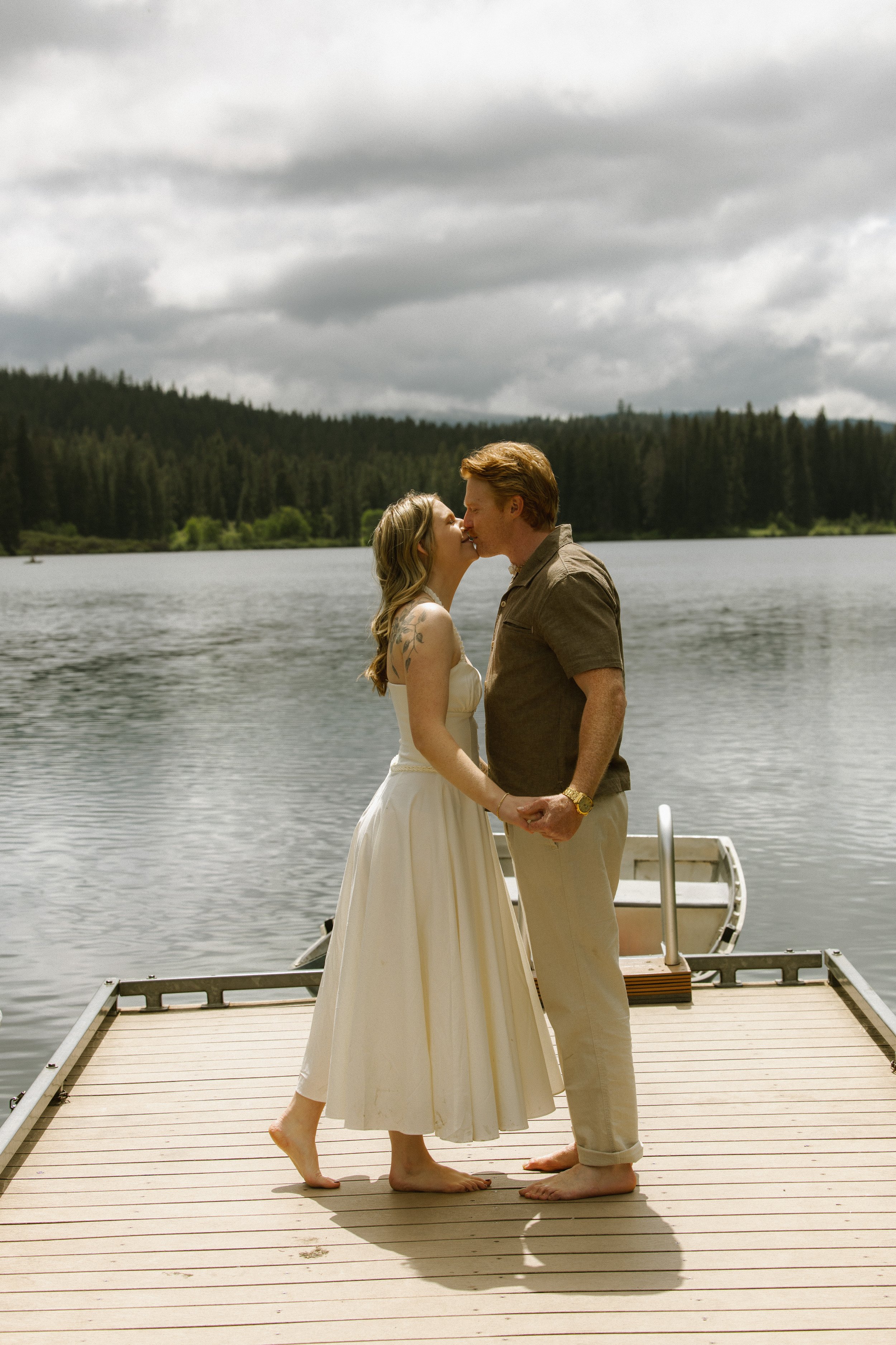 A couple stands on a dock by a lake, embracing and about to kiss, with a forested hillside under an overcast sky in the background.