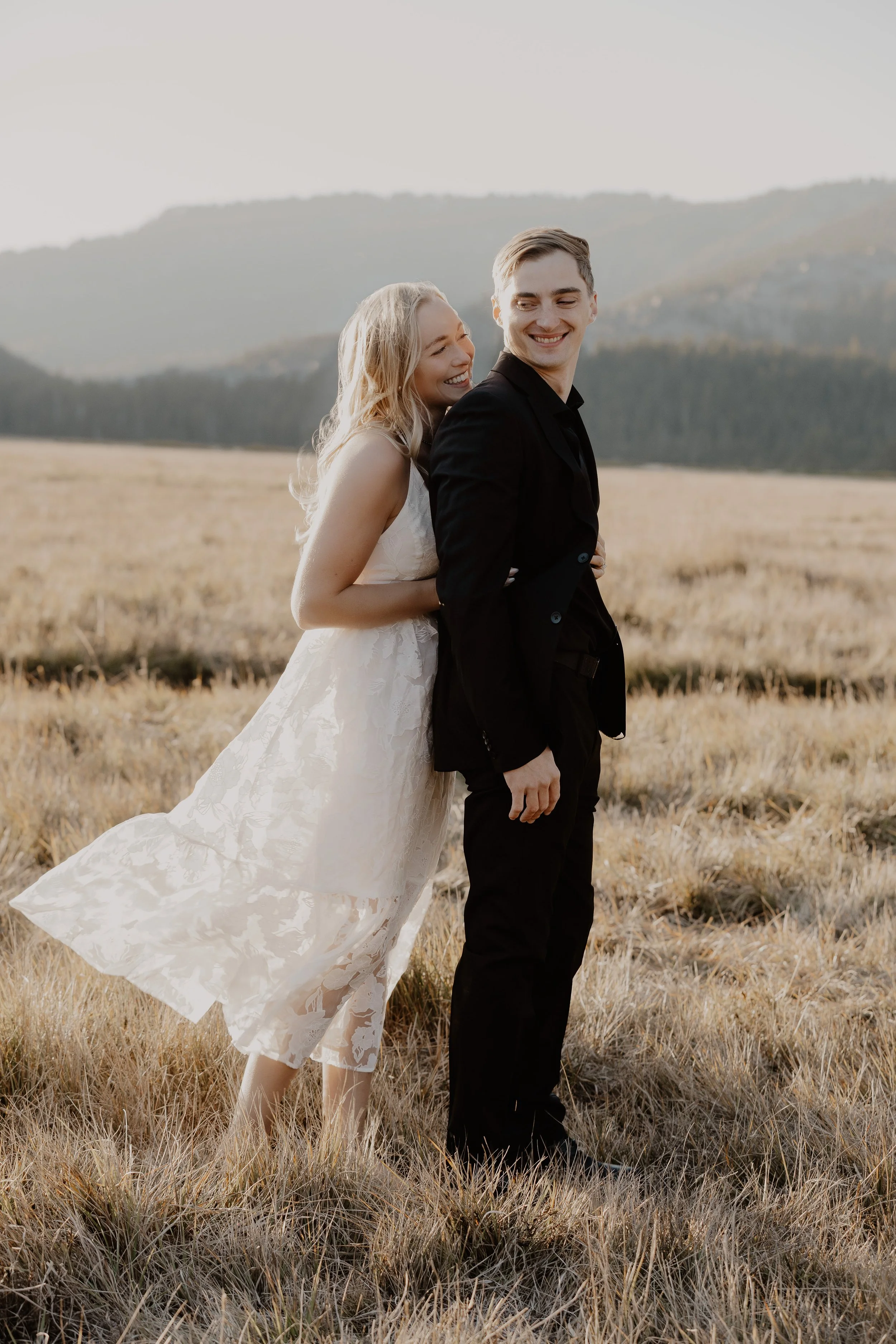 A couple in wedding attire standing in a field with mountains in the background, the woman in a white wedding dress and the man in a black suit, smiling and looking at each other.