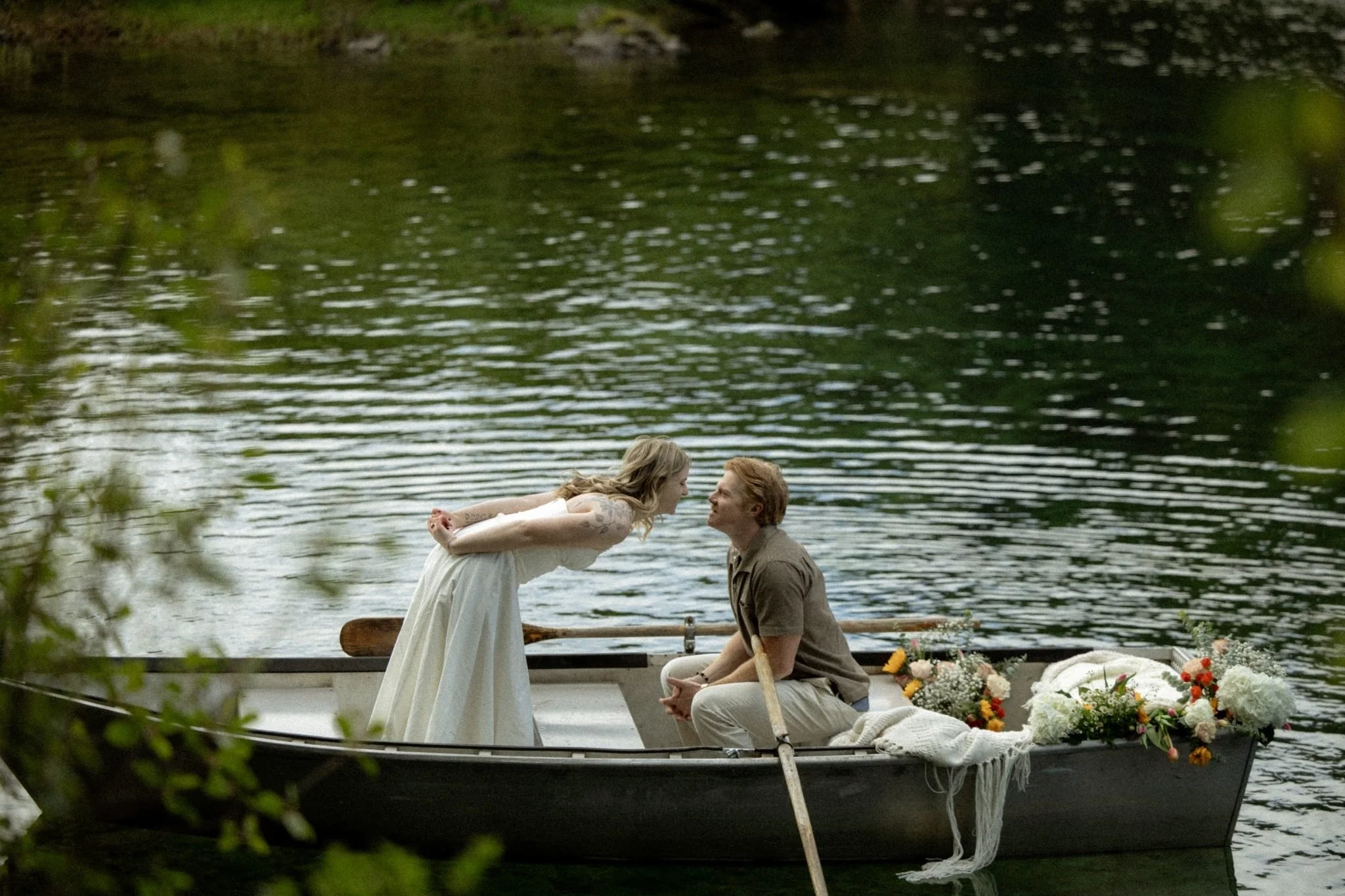 A woman in a white dress and a man in a brown shirt share a moment on a boat decorated with flowers by a lake, with trees and water in the background.