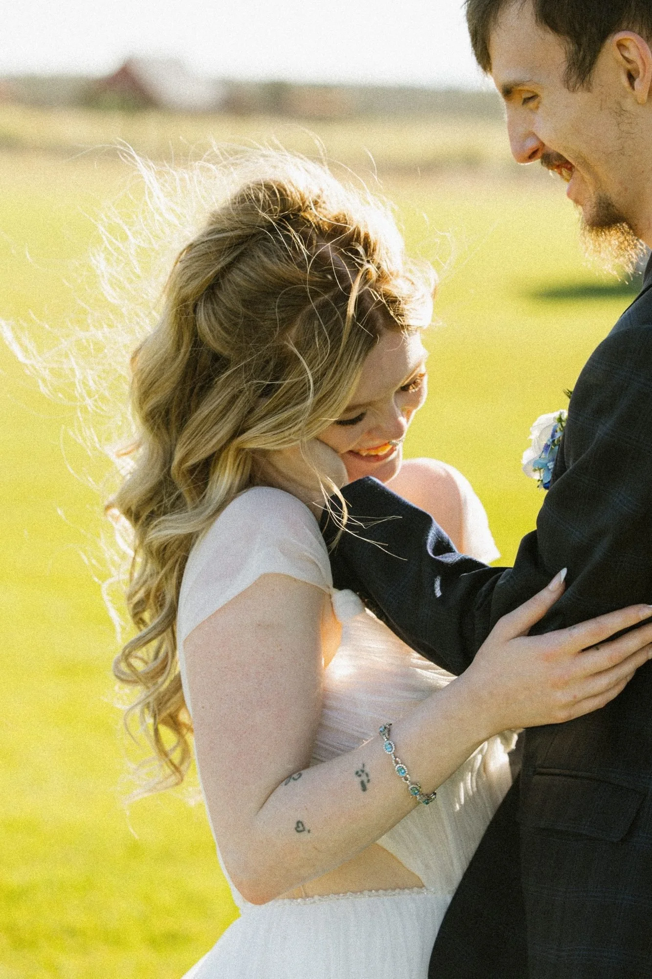 A couple smiling and embracing outdoors with a wedding cake in front of them on a table.