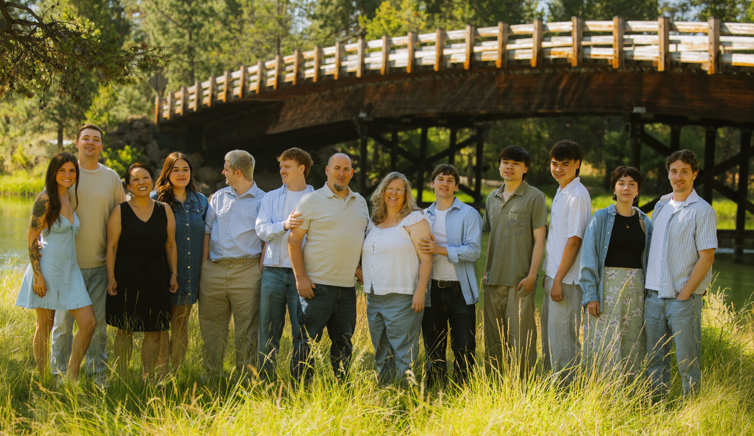 A group of fifteen diverse people standing outdoors near a body of water, with a wooden bridge and trees in the background, smiling at the camera.