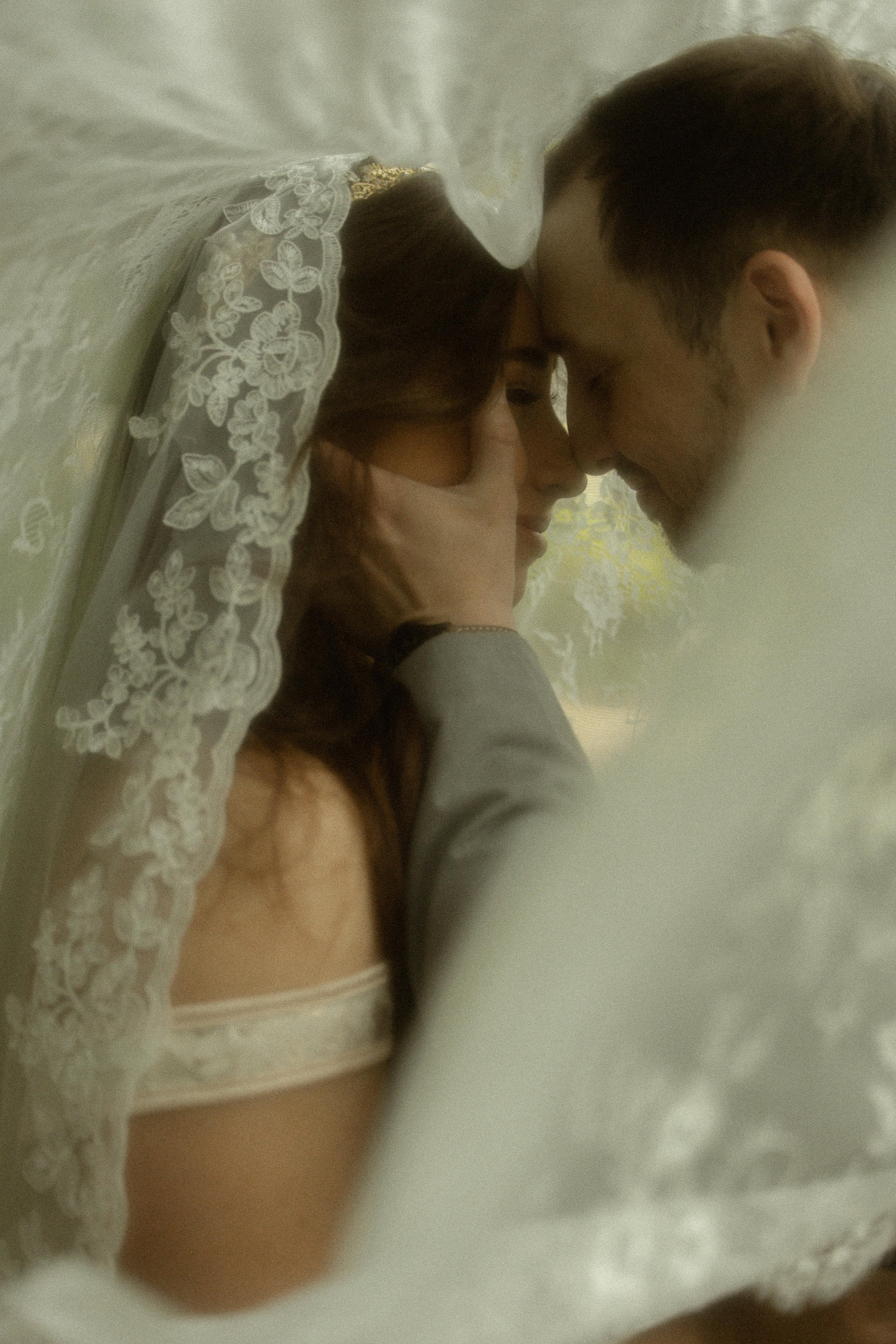 A couple, likely a bride and groom, in an intimate close-up moment, with their foreheads touching and eyes closed, dressed in wedding attire, with lace details visible.