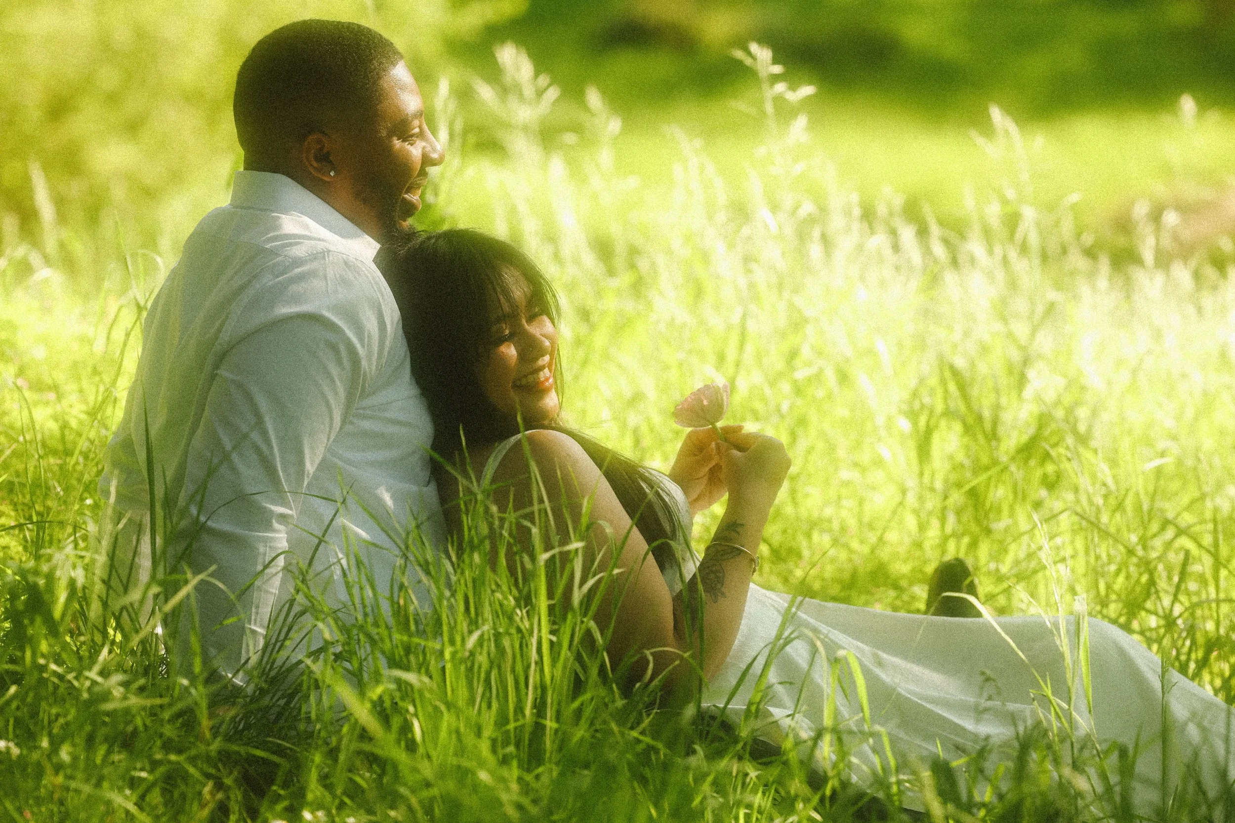 A smiling man and woman sitting in tall grass in a lush green field, enjoying nature on a sunny day.
