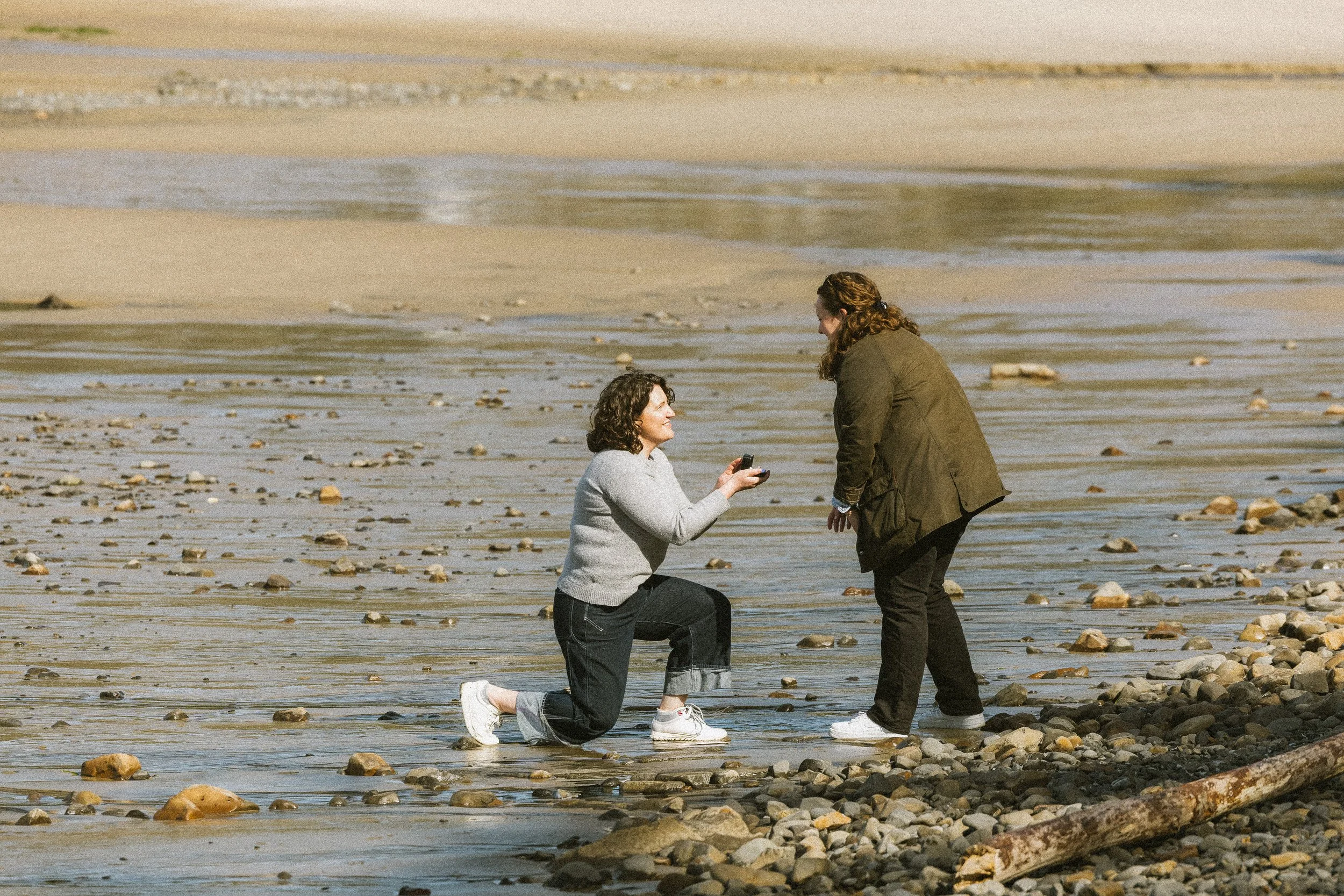 A woman kneeling on a rocky beach, proposing to a woman standing in front of her, with a scenic shoreline and water in the background.