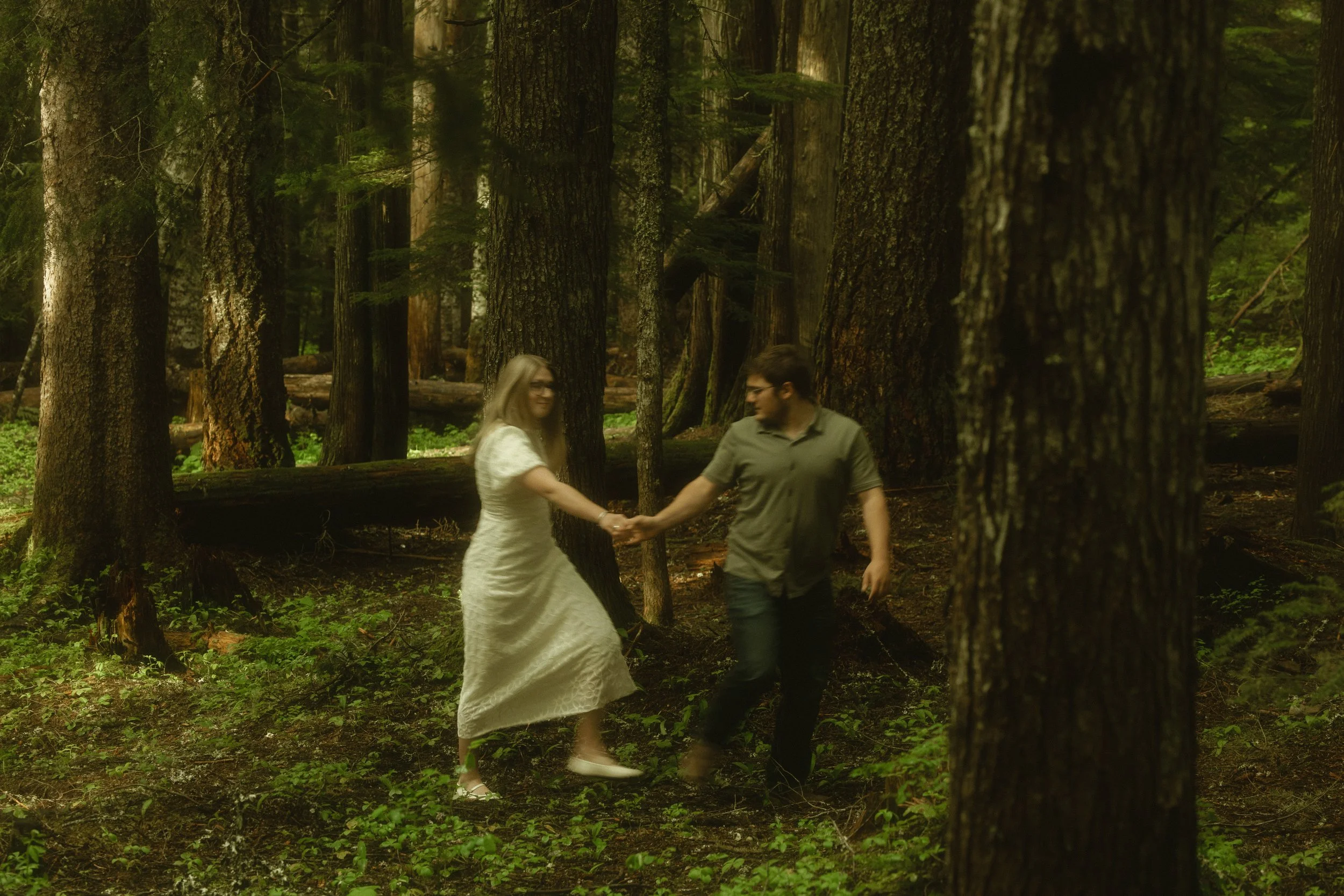 A woman in a white dress and a man in a gray shirt holding hands and walking in a dense forest with tall trees and green foliage.
