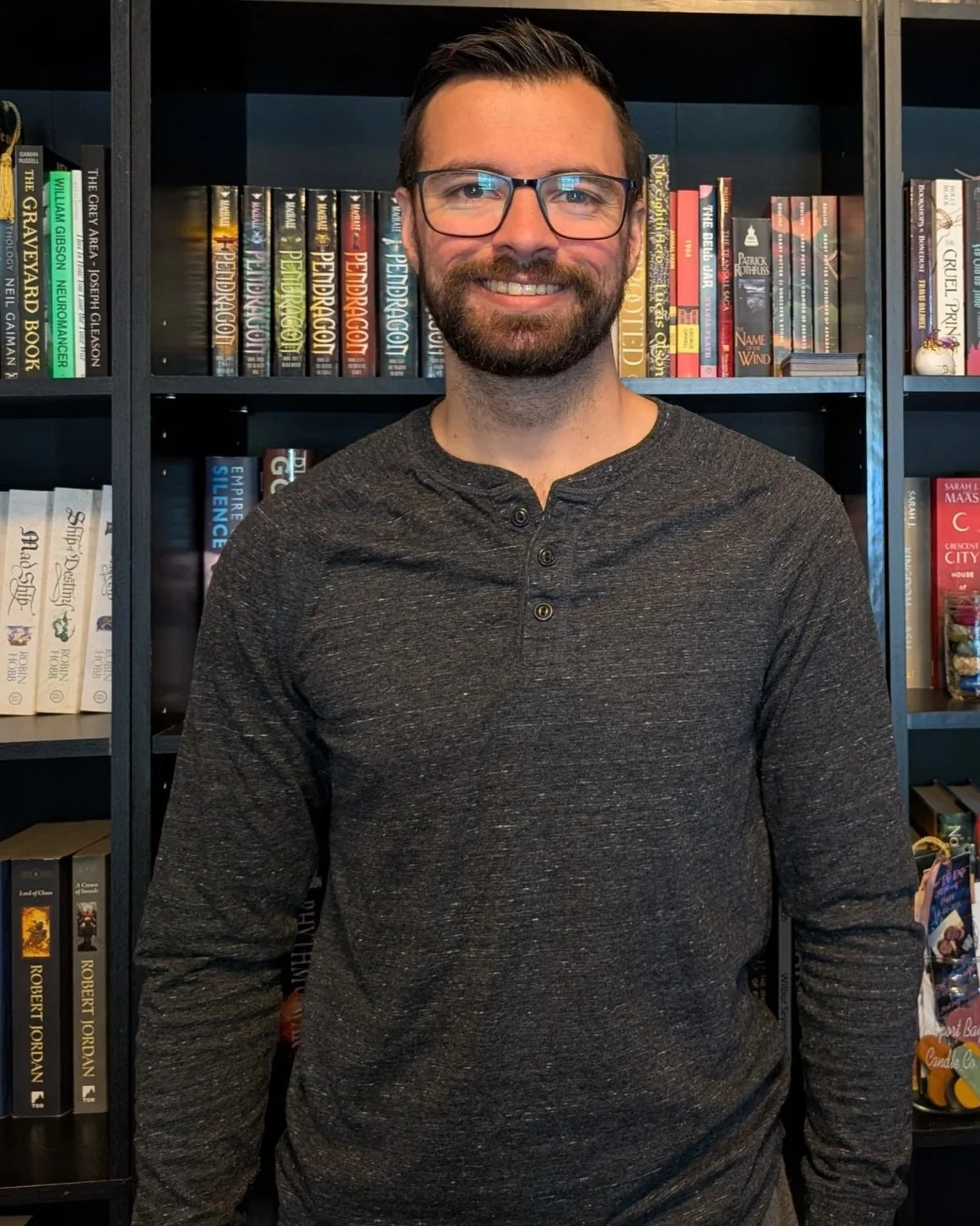 A smiling man with glasses, a beard, and short dark hair, wearing a dark gray long-sleeve shirt, stands in front of a bookshelf filled with books.