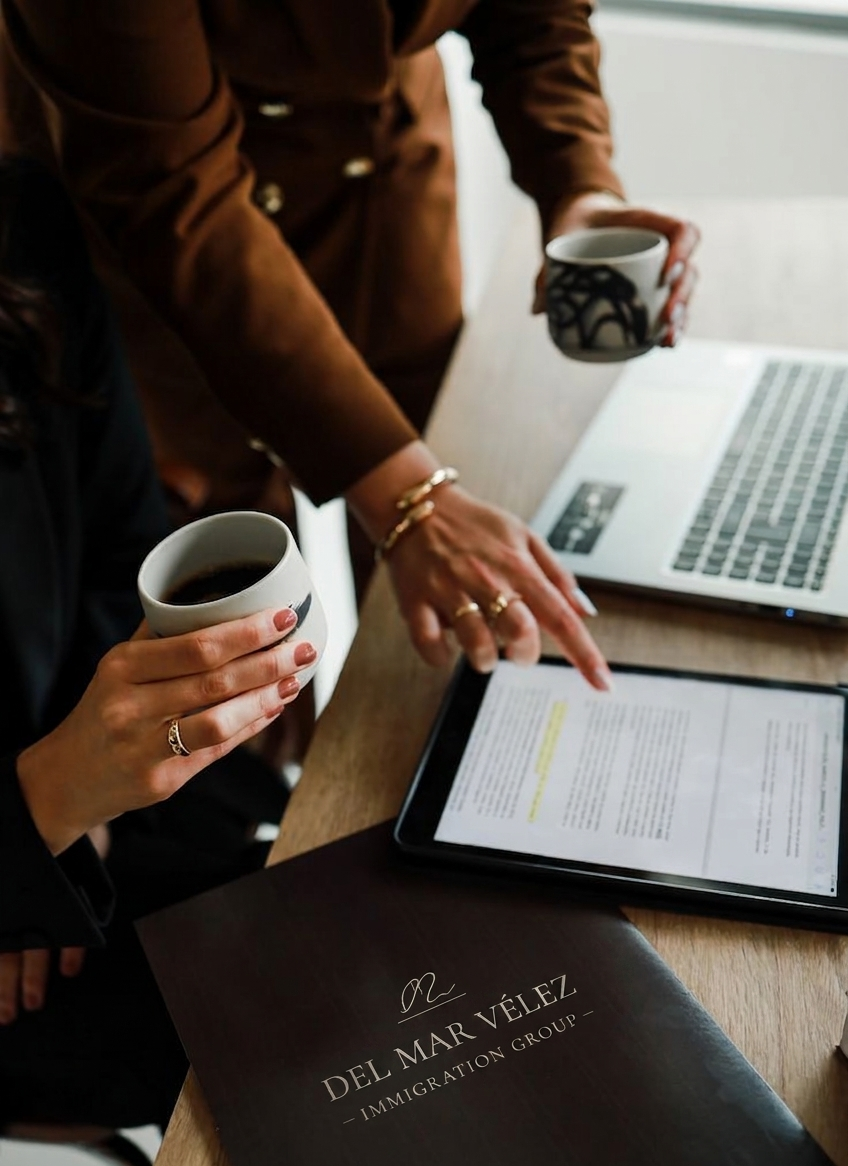 Two women are sitting at a table with a laptop, an iPad, and a brochure from Del Mar Velez Immigration Group. They are holding coffee mugs, and one woman is pointing at the iPad screen.